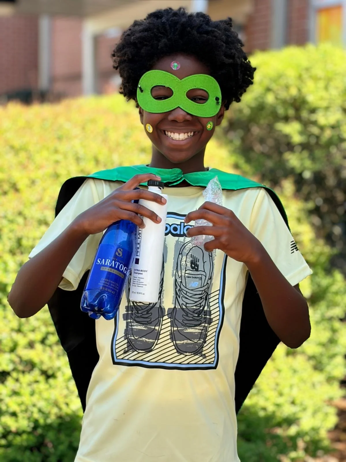 A smiling young child dressed as a superhero with a green mask, a yellow shirt with a skeleton graphic, holding shampoo and body wash, standing outdoors against a blurred background of greenery and buildings.