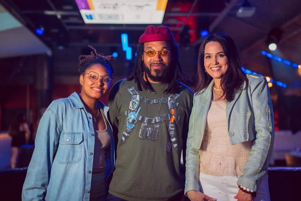 Three people smiling together in a dimly lit indoor space with colorful lights, one woman with glasses and dreadlocks, one man with dreadlocks and wearing a red beanie, and another woman with dark hair and a shiny jacket.