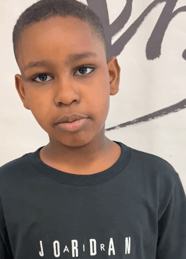 Close-up of a young boy with short black hair, brown skin, and wearing a black shirt, standing in front of a white wall with a black abstract design.