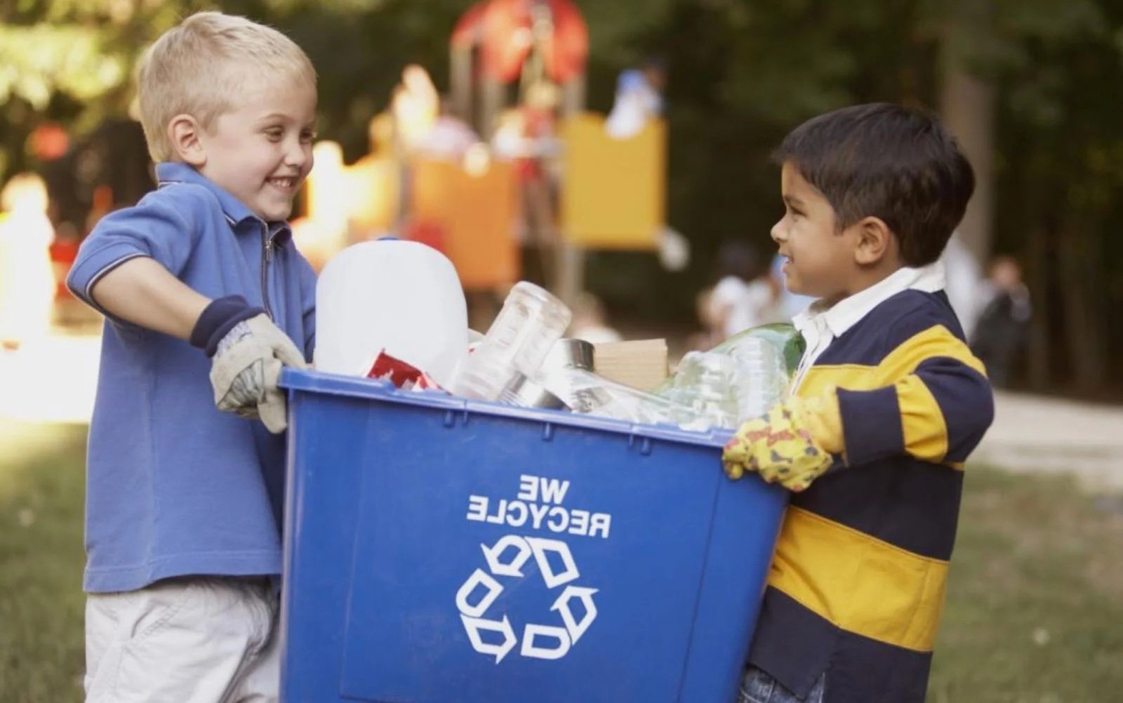 Two boys smiling and holding a large blue recycling bin filled with empty glass bottles and other recyclables at a park.