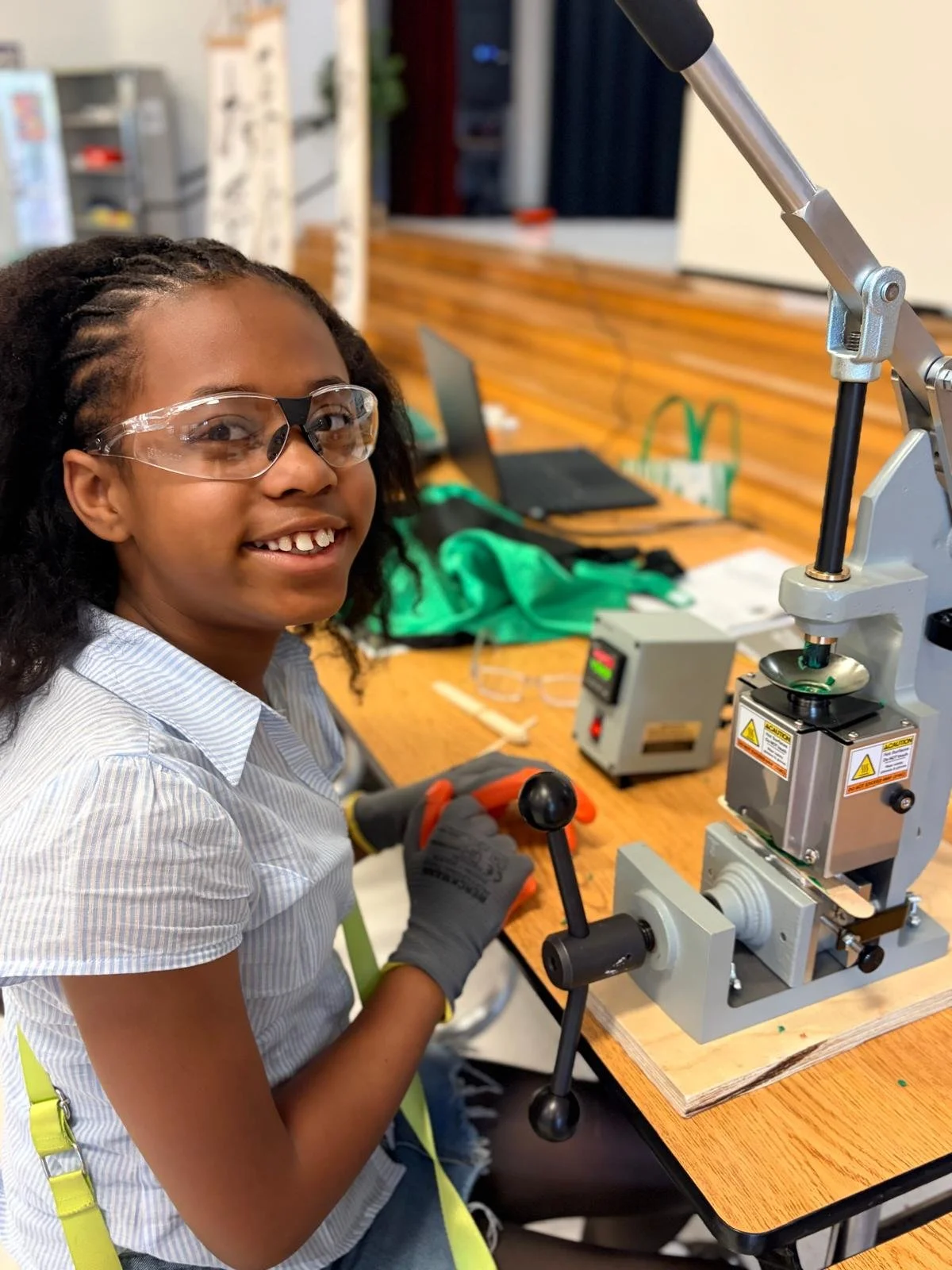 A young girl wearing safety goggles and gloves working with a scientific instrument at a lab table in a classroom or laboratory setting.