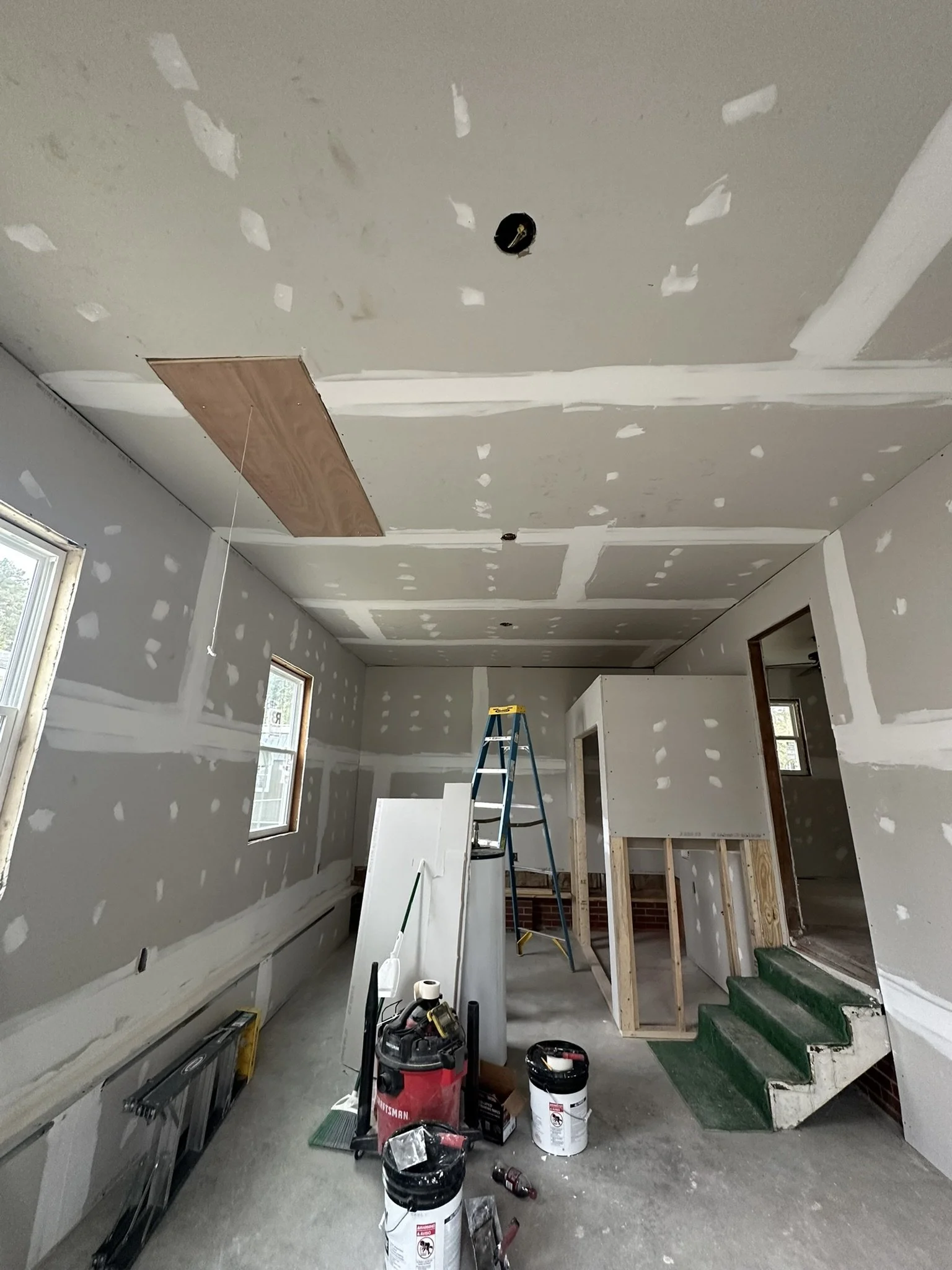 Interior of a house under construction with drywall, a ladder, stairs with green carpet, tools, and supplies.