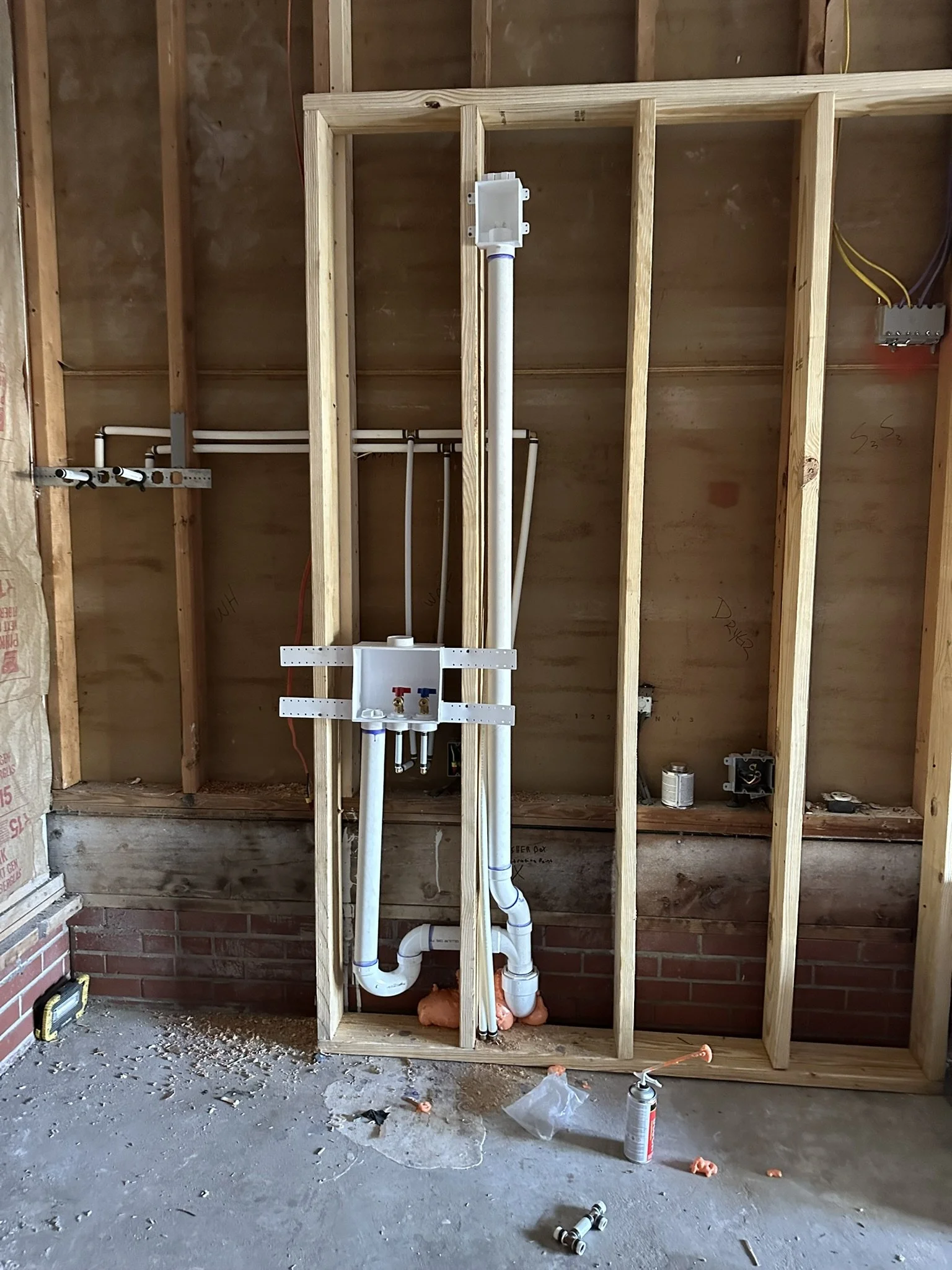 Open wall with exposed wooden studs, plumbing pipes, and electrical wiring in a construction site.