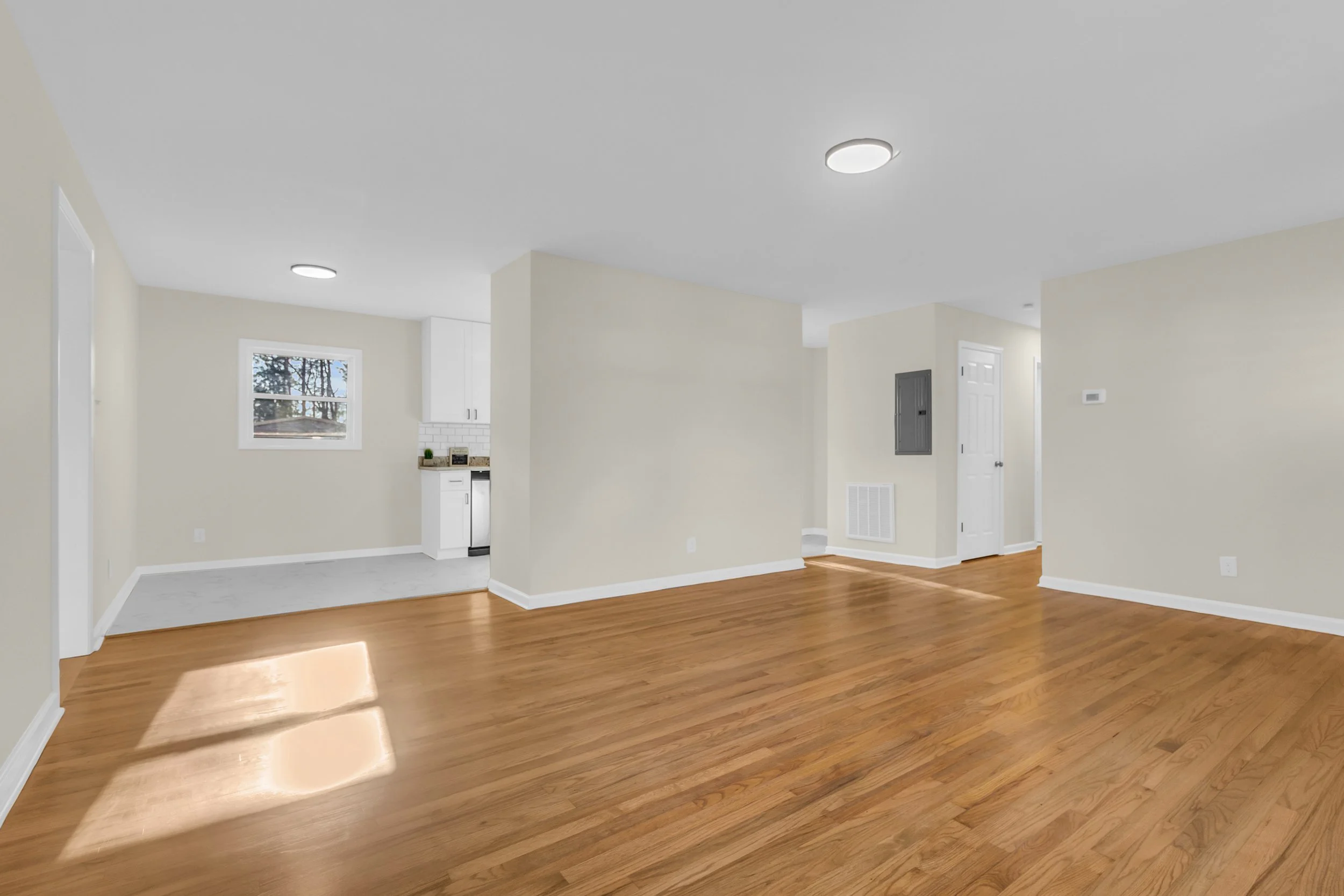 Empty living room with hardwood floors, white walls, and ceiling lights, adjacent to a kitchen area with white cabinets and a small window.