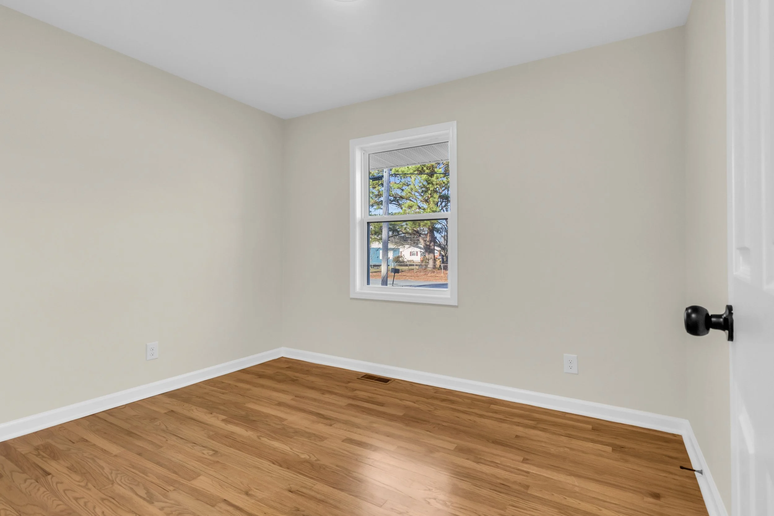 Empty room with beige walls, a window showing trees outside, hardwood floor, and a white door with a black doorknob.