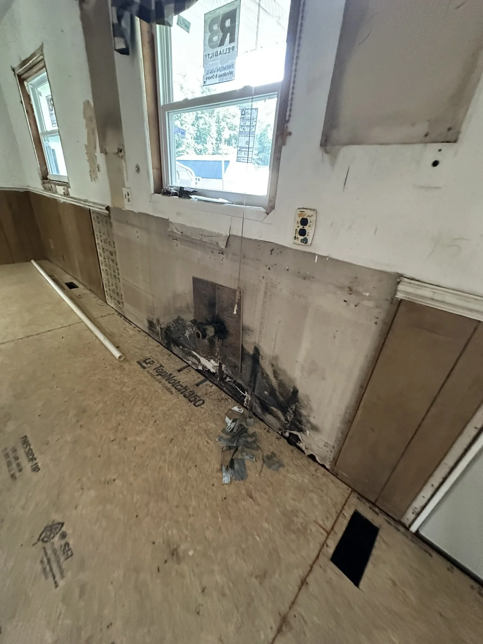 Partially demolished kitchen wall with exposed electrical outlet and debris on the floor, near window.