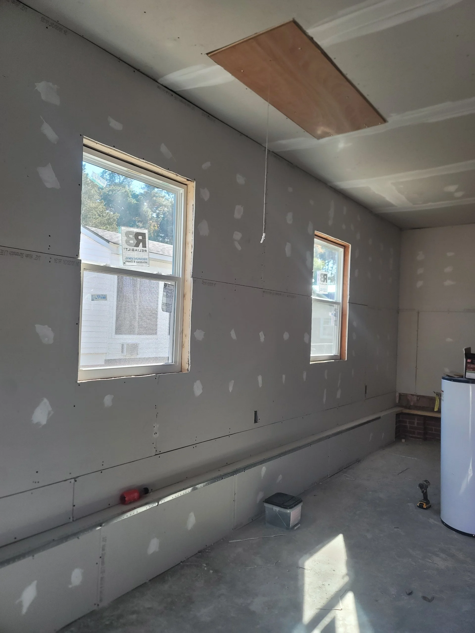 Interior of a room under construction with partially finished drywall, two windows, a ceiling hatch, and construction tools on the floor.