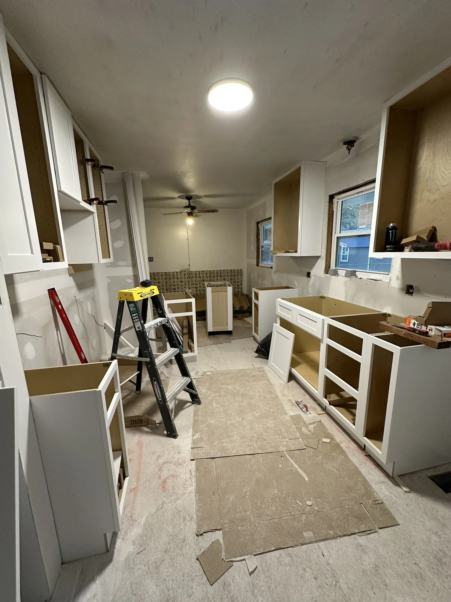 Kitchen under renovation with white cabinets, some installed and some in progress, and construction tools like a step ladder and level scattered around.