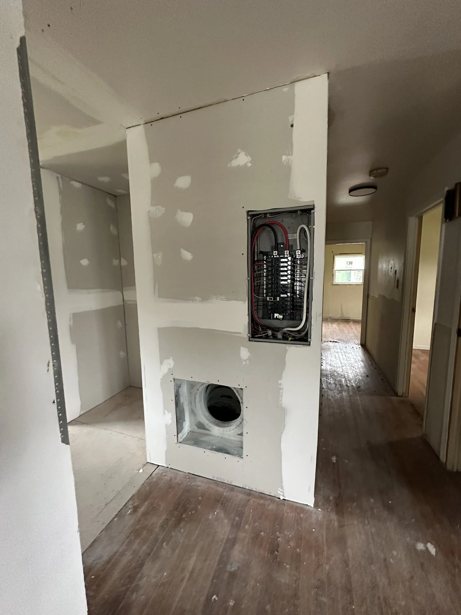 Interior view of a house under renovation, showing an unfinished wall with electrical panel and pipe opening, with rooms in the background.