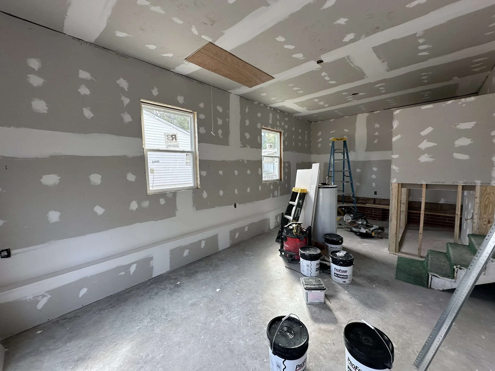 An unfinished room under construction with drywall, windows, and carpentry tools, buckets of paint, a stepladder, and stairs.
