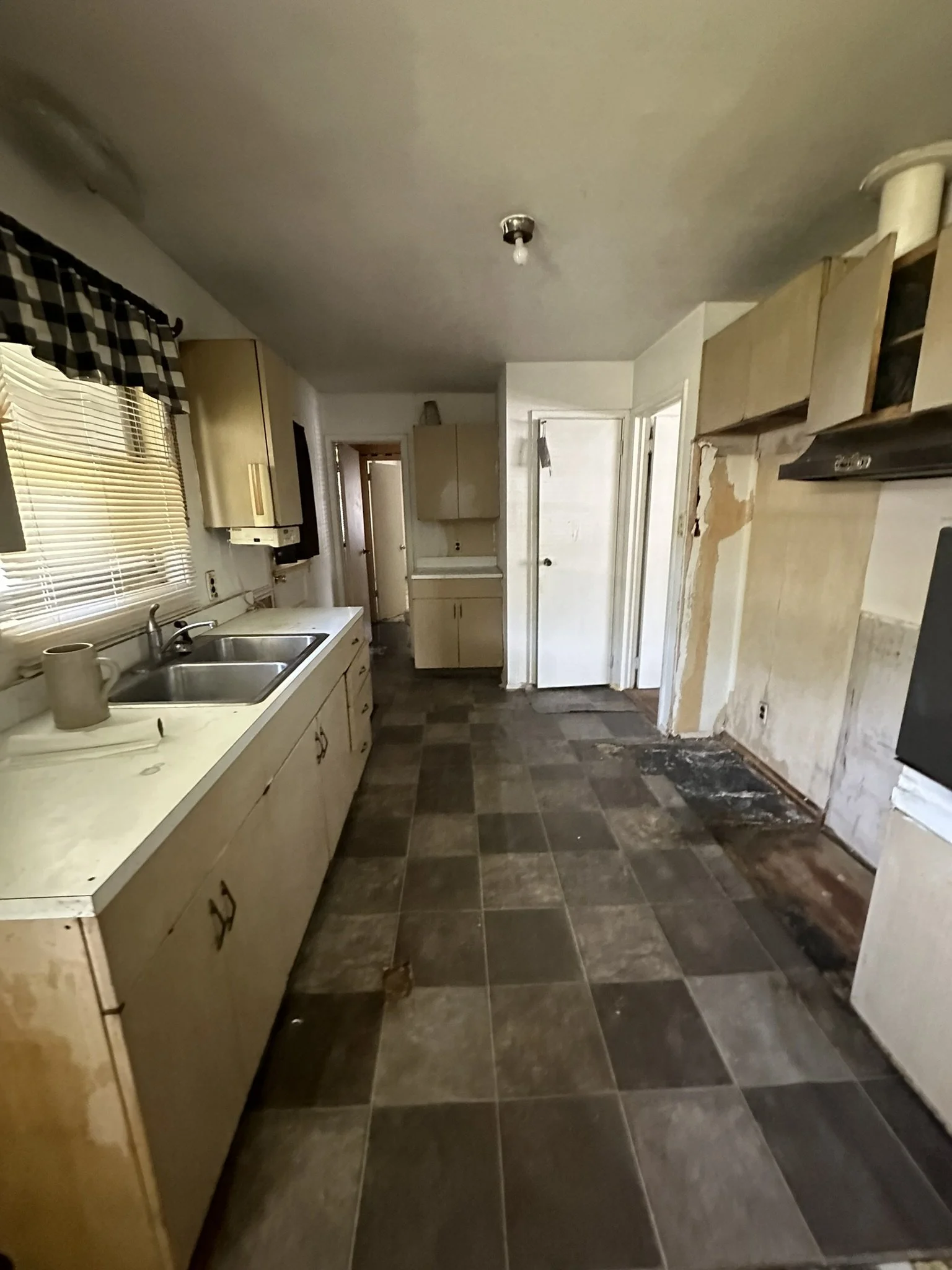A kitchen with old cabinets, a double sink, window with blinds and checkered valance, and visible signs of renovations with missing cabinet doors and damaged flooring.