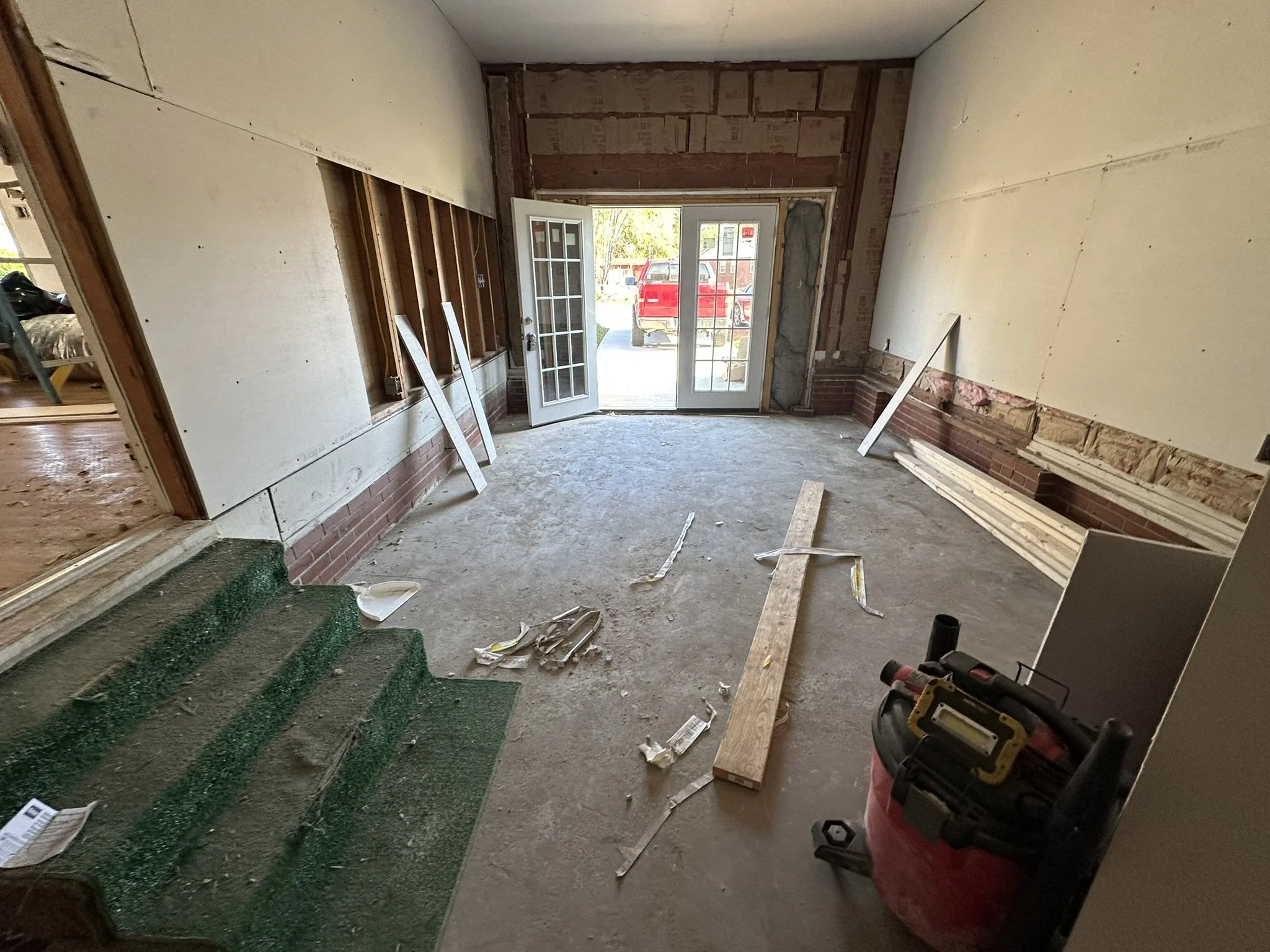Room under renovation with exposed brick and drywall, some construction materials on the floor, and an open front door leading outside.