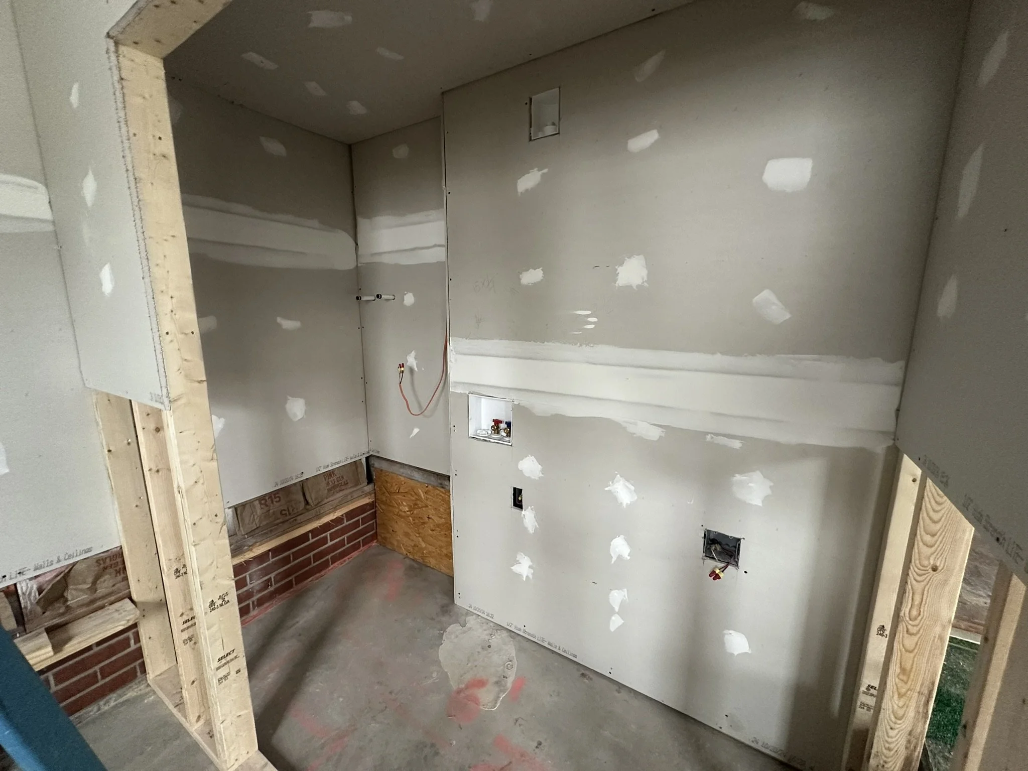 Interior of a room under construction with drywall installed, unfinished walls, exposed electrical outlets, and framing. The walls have patches of drywall mud, and the floor is bare concrete.