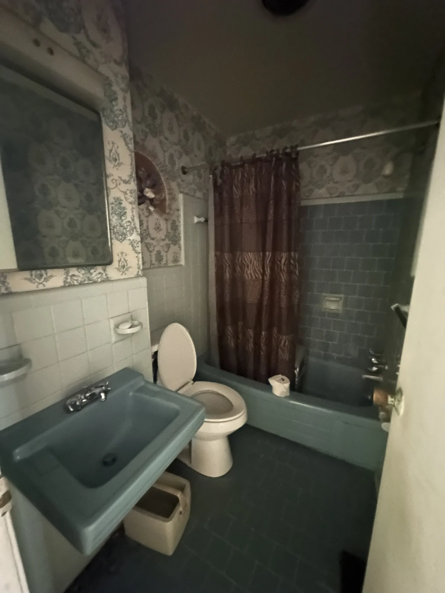 A small vintage bathroom with teal fixtures, including a sink, toilet, and bathtub, with patterned wallpaper and a dark shower curtain.