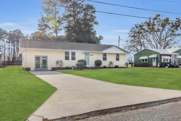 Front view of a single-story yellow house with white trim and a concrete driveway, surrounded by a well-maintained lawn and trees in the background.