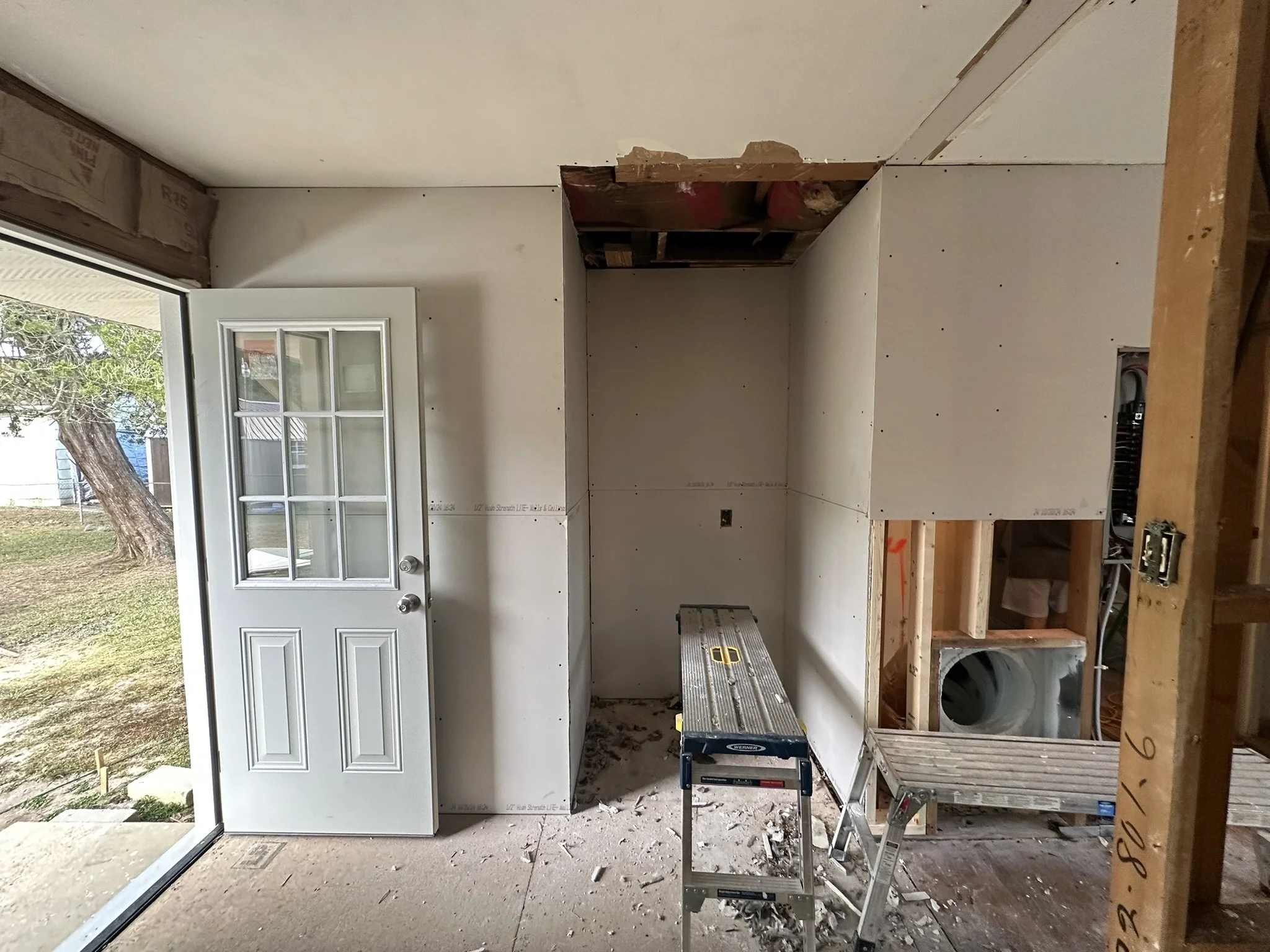 Room under renovation with drywall installation, a glass door open to outside, construction tools, and material debris.