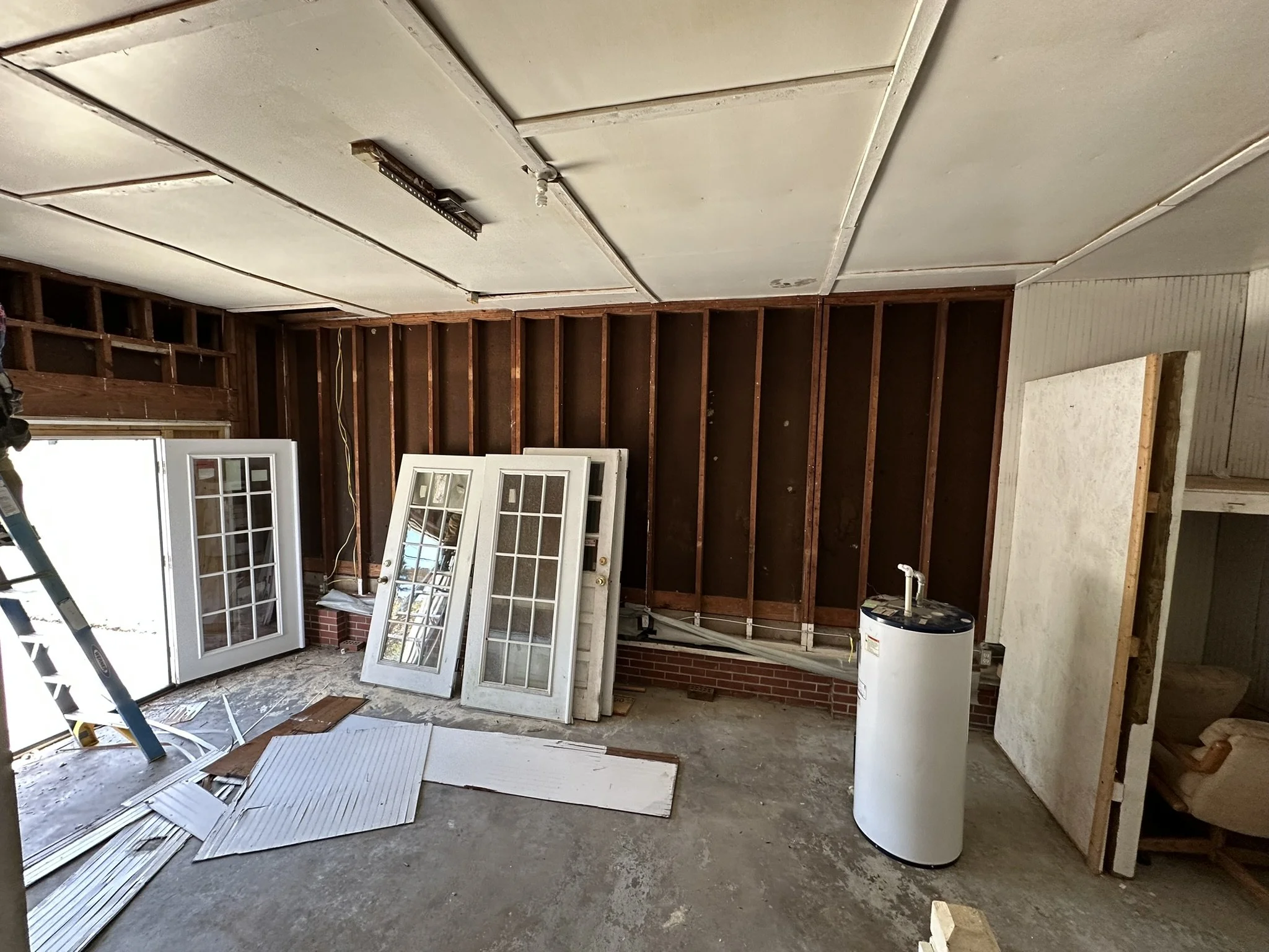 Room under renovation with exposed wooden wall studs, some detached glass door panels on the floor, a ladder, a water heater, and construction debris.