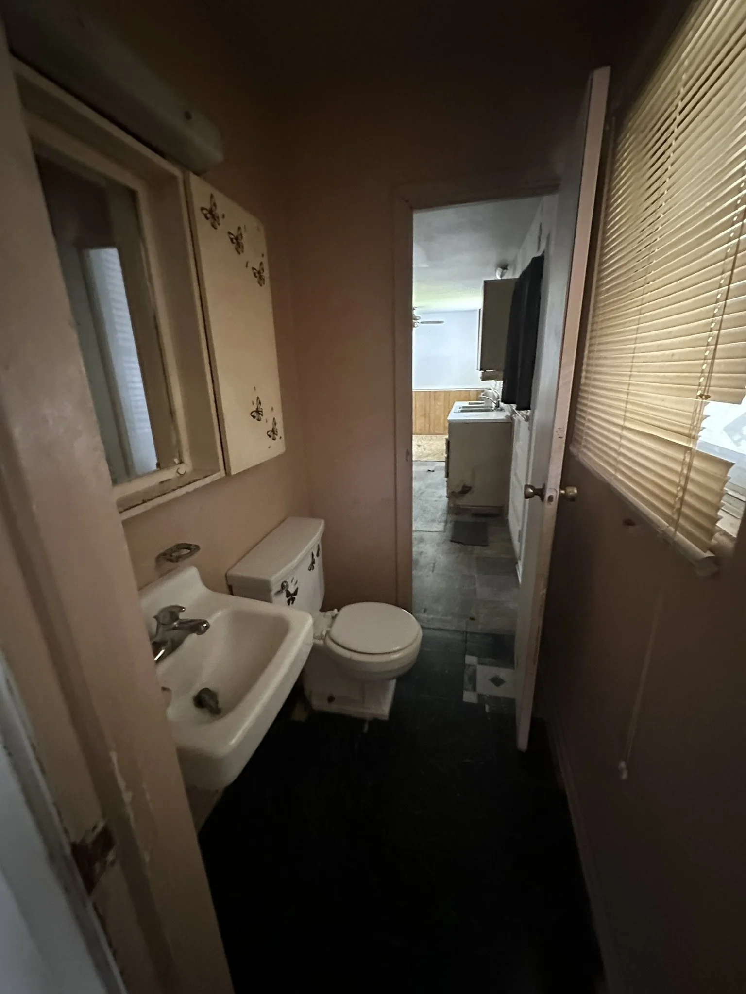 A small bathroom with a toilet, a sink, a window with closed blinds, and a mirror with butterfly stickers on the wall. The doorway leads to a room with a washing machine and a TV.