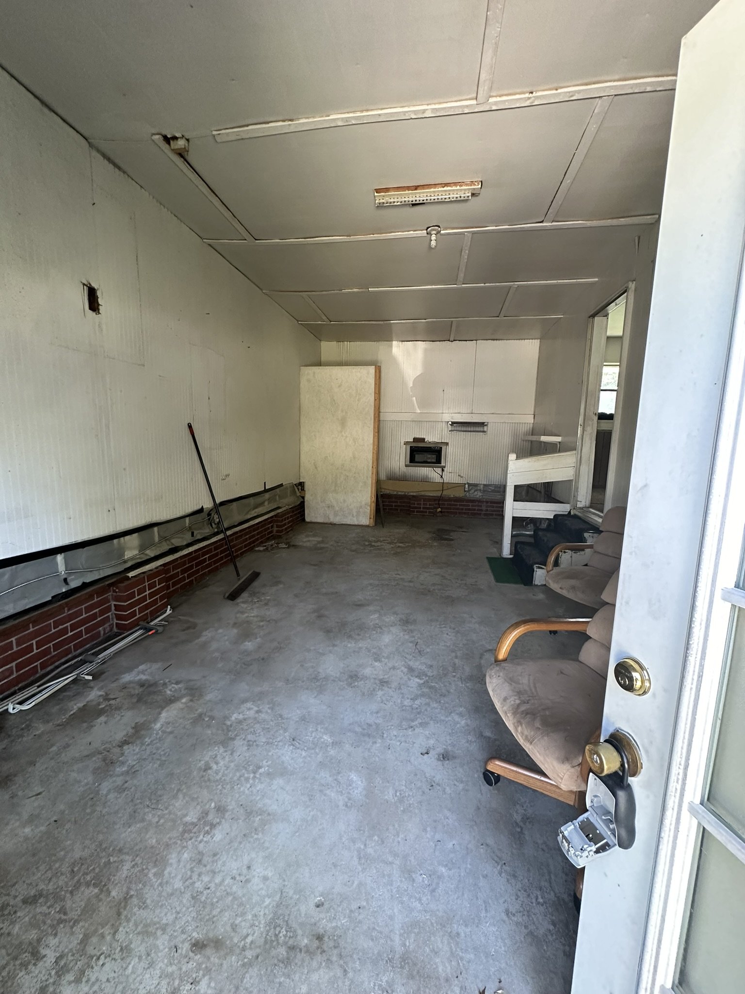 Empty porch with concrete floor, brick and panel walls, a brown armchair, and some miscellaneous items leaning against the wall.