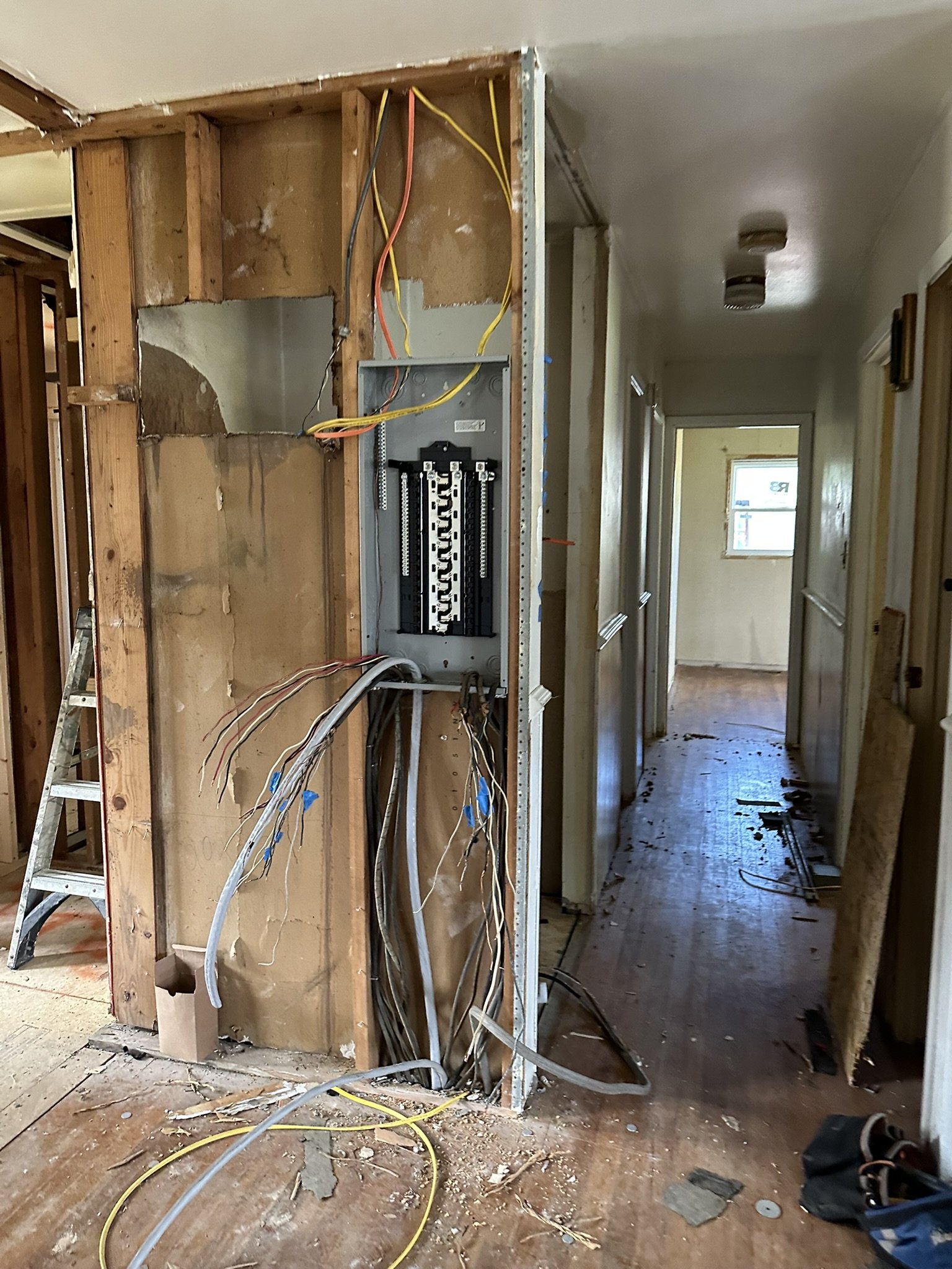 Open wall with electrical wiring and a breaker box under renovation in a hallway of a house.