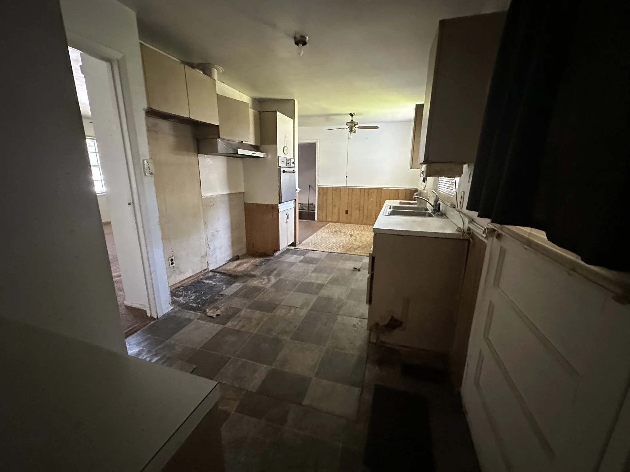A kitchen in need of renovation with worn wooden cabinets, a mixed tile floor, and a white double sink. The window has black curtains. The room opens into a dining area with a ceiling fan and wood-paneled walls.