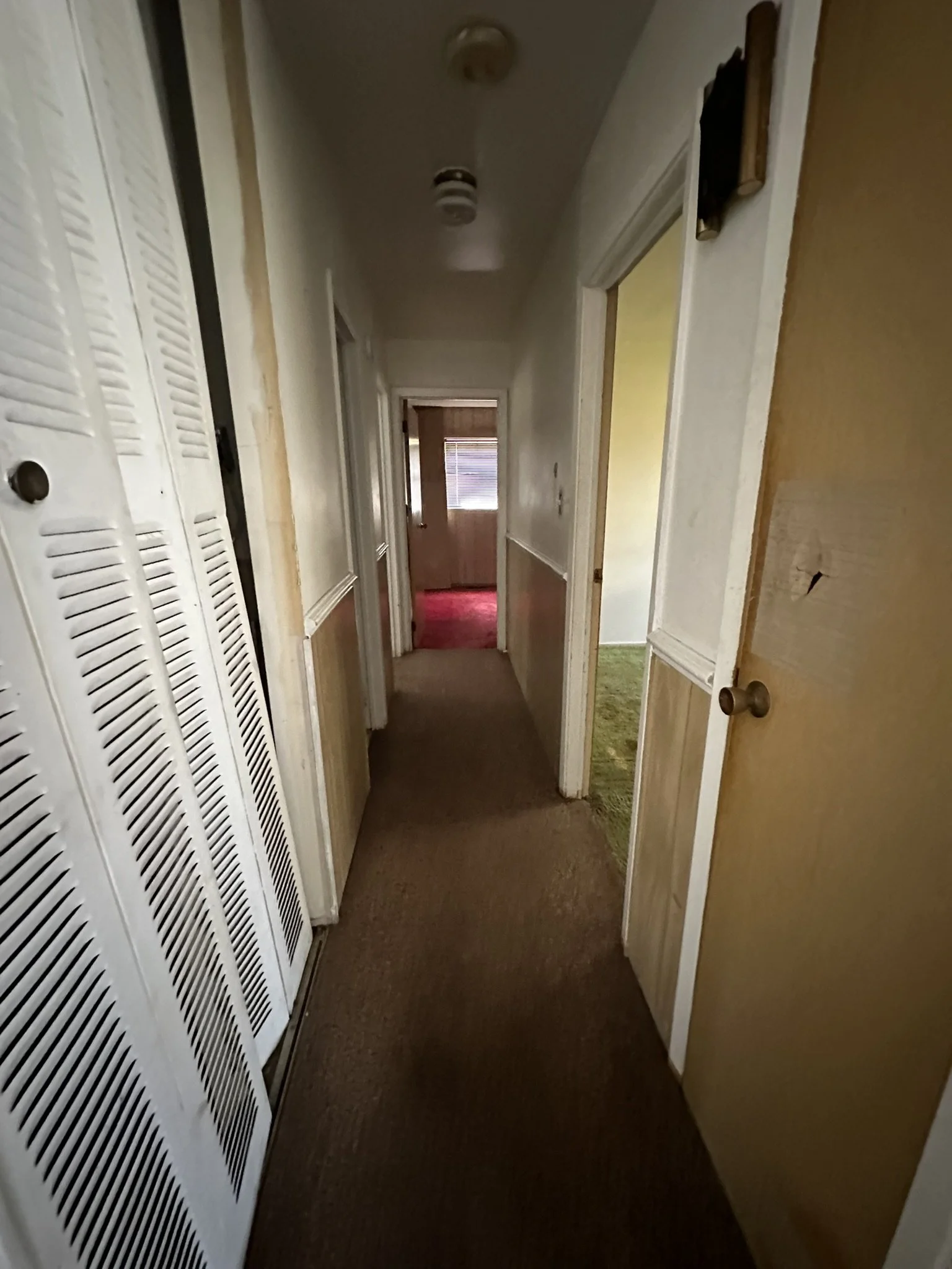 A narrow indoor hallway with brown carpet, beige walls, and white door frames. Doors on the right, some slightly open, show rooms with green and yellow carpets. A closet with louvered white doors is on the left. The hallway has a ceiling light fixtur