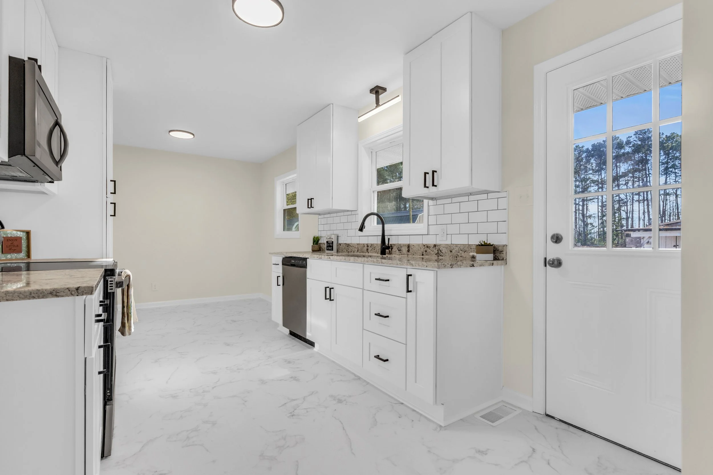 Modern kitchen with white cabinets, granite countertops, stainless steel appliances, white subway tile backsplash, and a door with a window leading outside. Natural light coming through two windows.