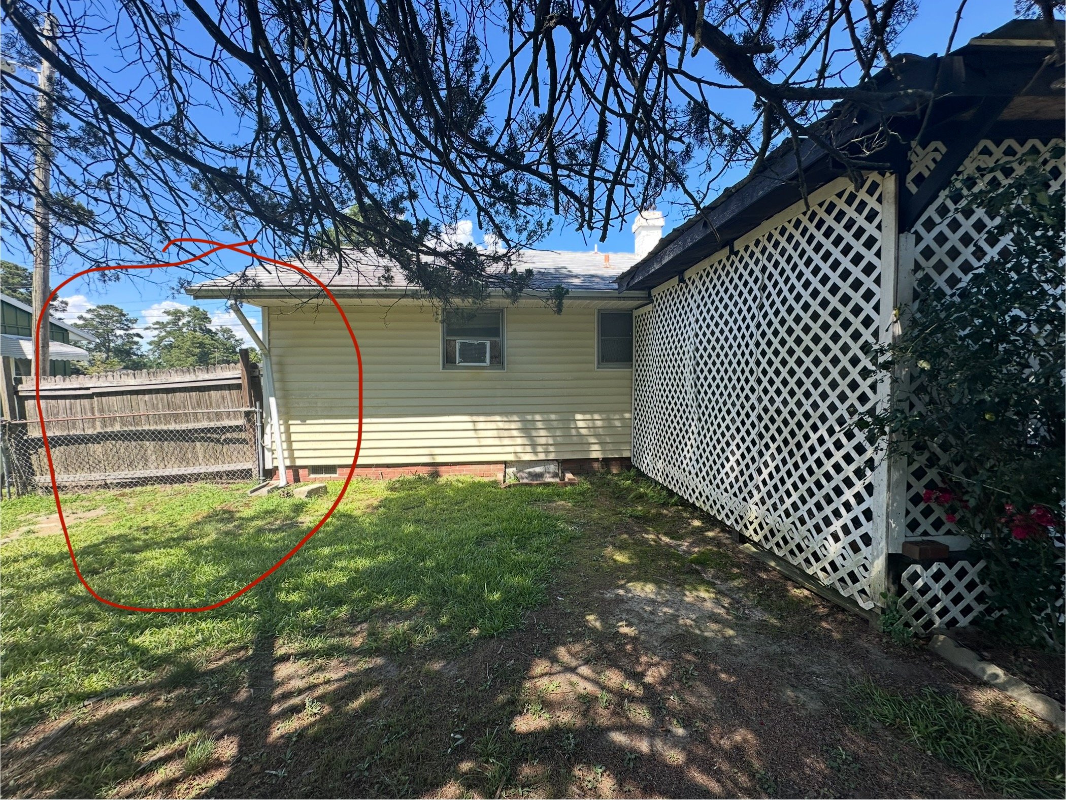Residential backyard with grass, trees, and fences. There is a house with beige siding, two small windows, and a white lattice structure attached to the house. The sky is blue with some clouds.