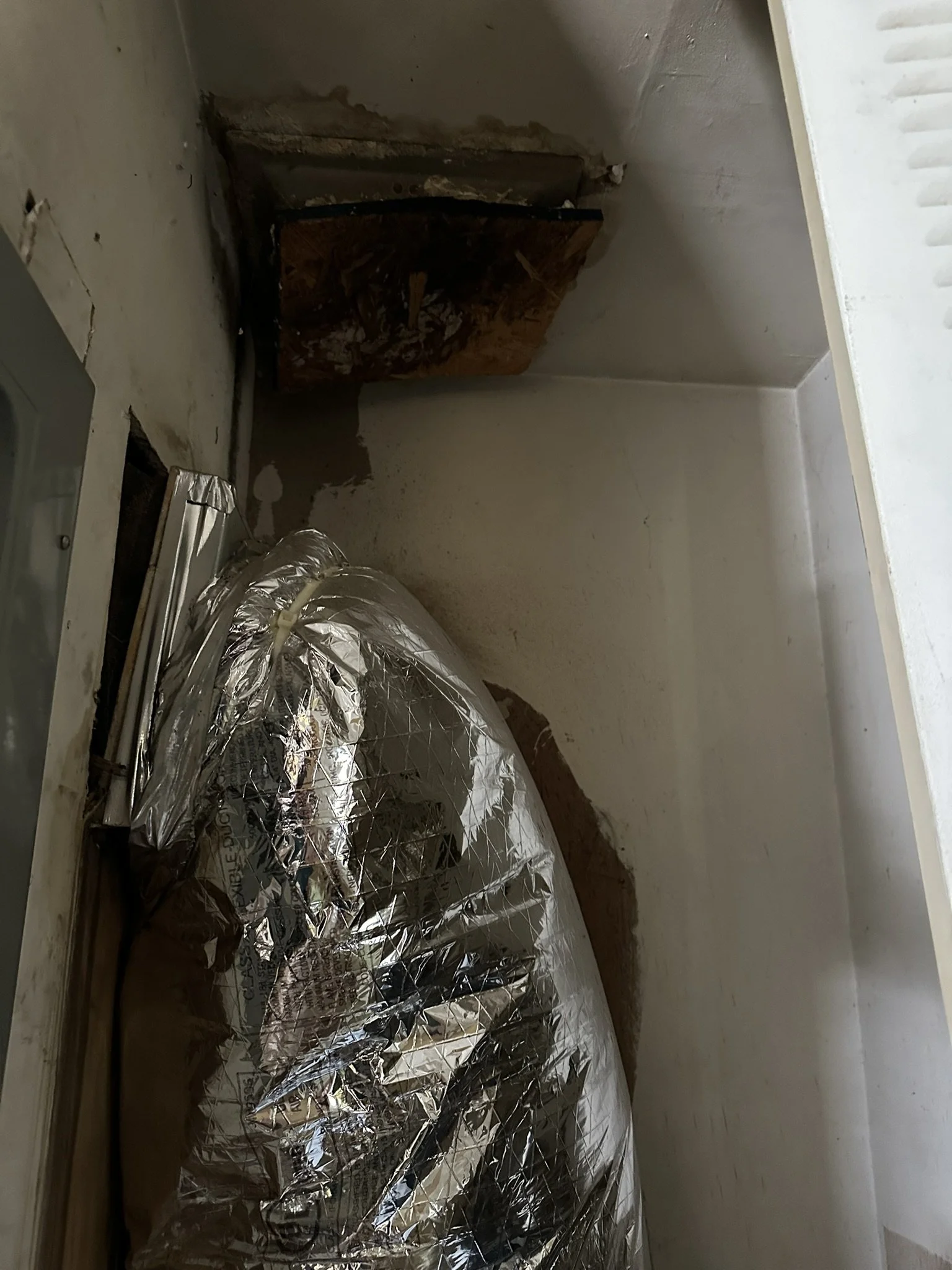 A space under a kitchen sink with an open cabinet door. The area is damaged and has a piece of wood blocking the opening. There is a large object wrapped in reflective insulation material resting on the floor near the cabinet.