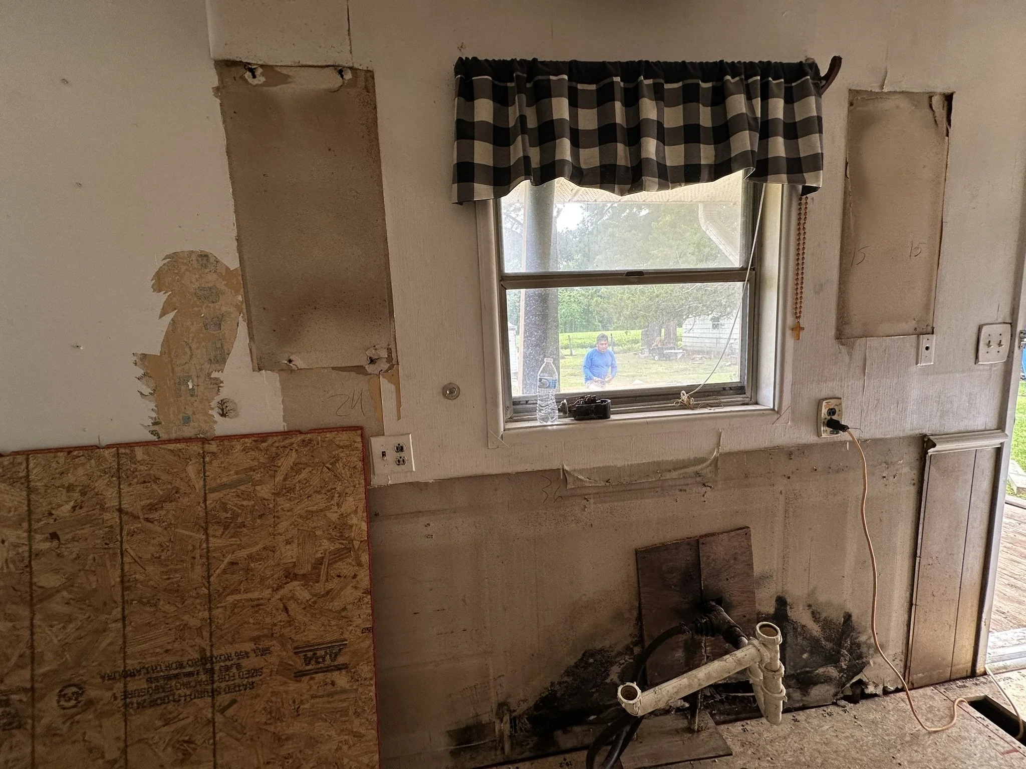 A partially renovated kitchen or utility room with exposed drywall, an empty wall cavity, and plumbing pipes. There is a window with a black and white checkered valance, looking outside at a person in a blue shirt and green outdoor setting. The wall 