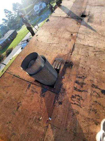A black vent pipe protruding from a wooden roof or deck, with workers and trees visible in the background.