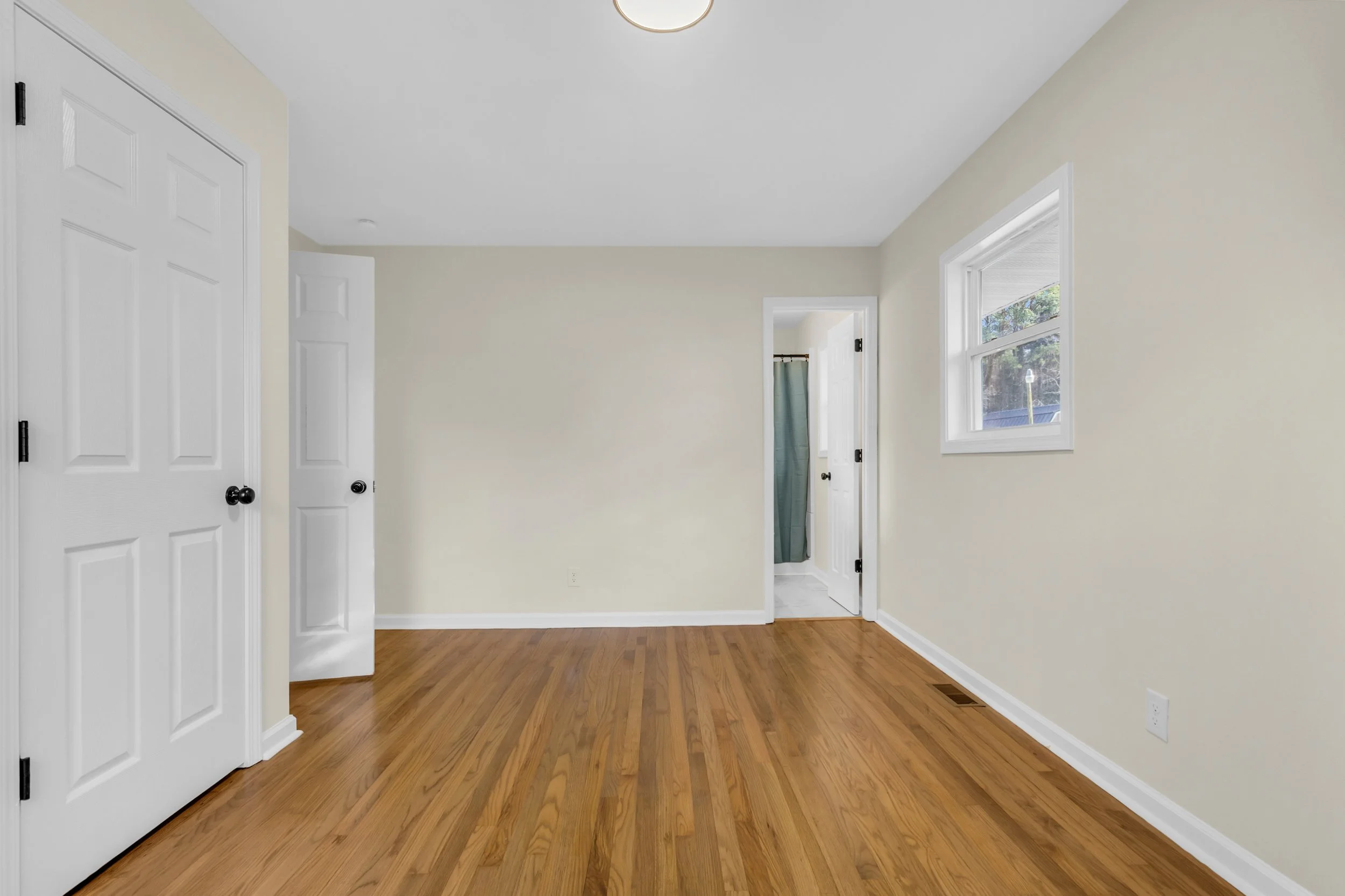 Empty room with hardwood floors, cream walls, a window, two closed closet doors, and an open door leading to a bathroom with a teal shower curtain.