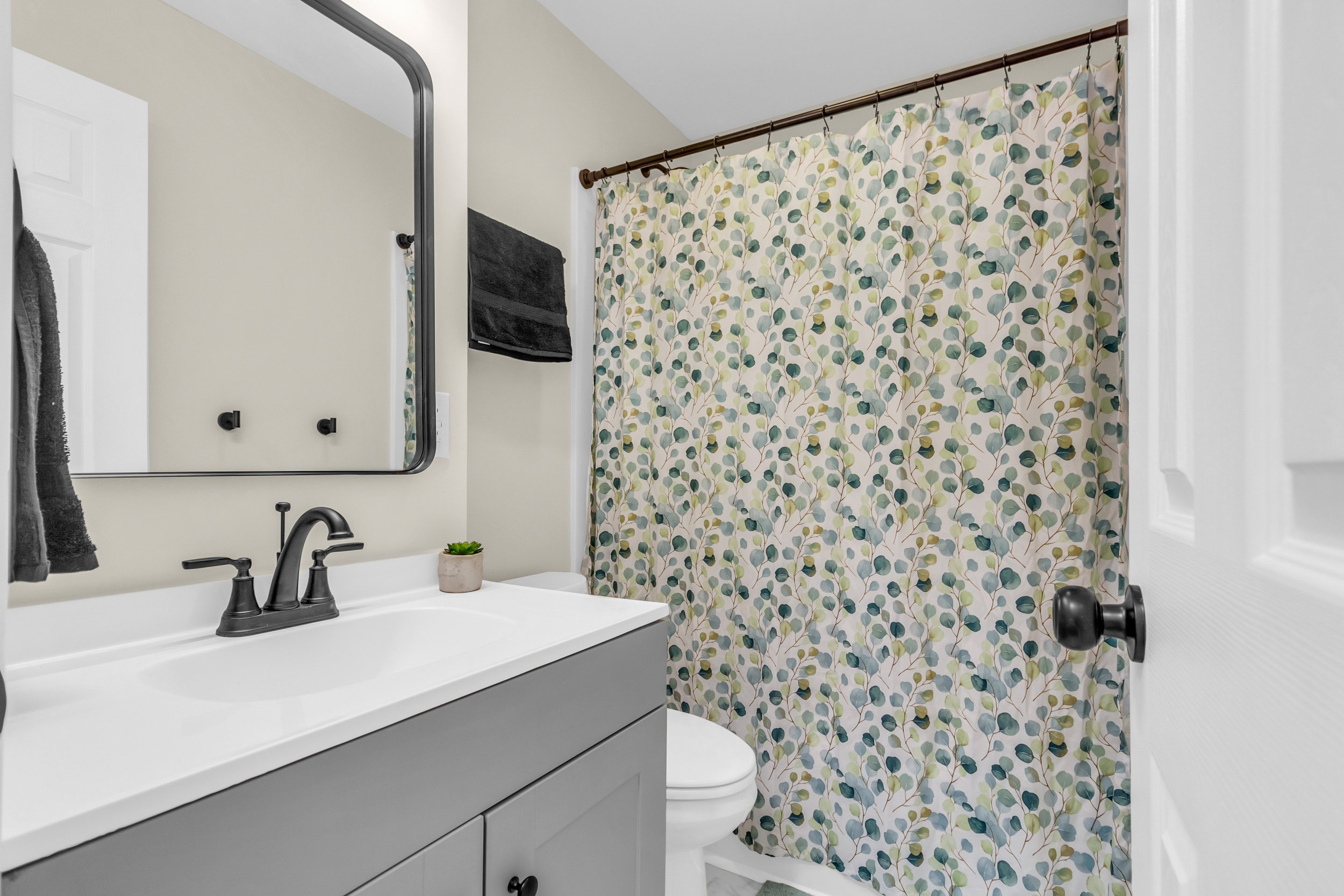 Bathroom with a gray vanity, white sink, black faucet, decorative shower curtain with leaf pattern, mirror, black towel, and small plant on the counter.
