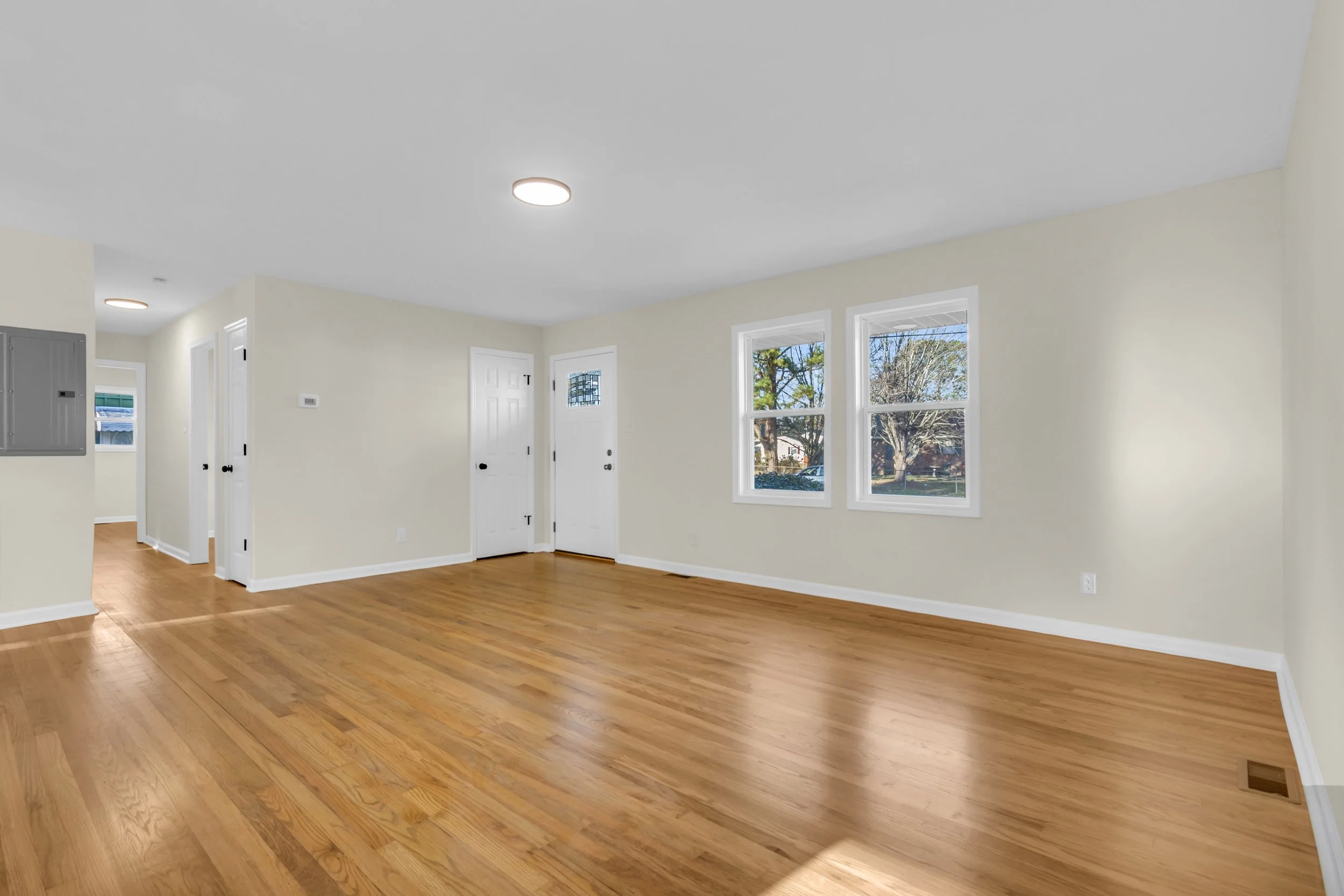 Empty living room with hardwood floors, beige walls, and three large windows showing trees outside.
