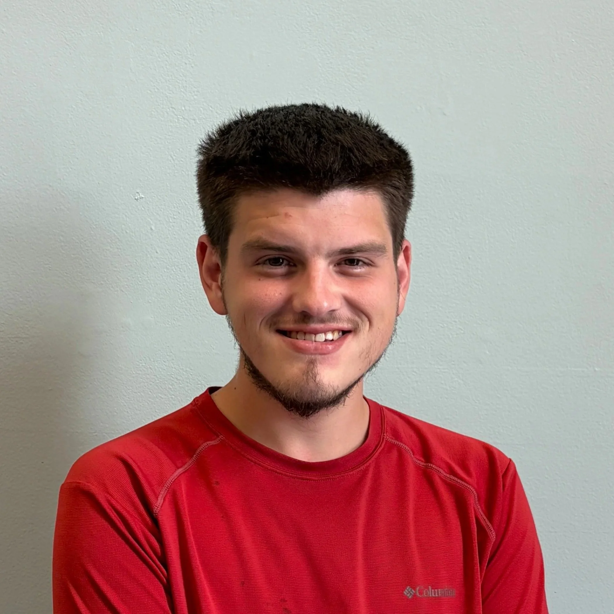 Close-up of a smiling man wearing a red Columbia shirt with a plain light-colored wall in the background.