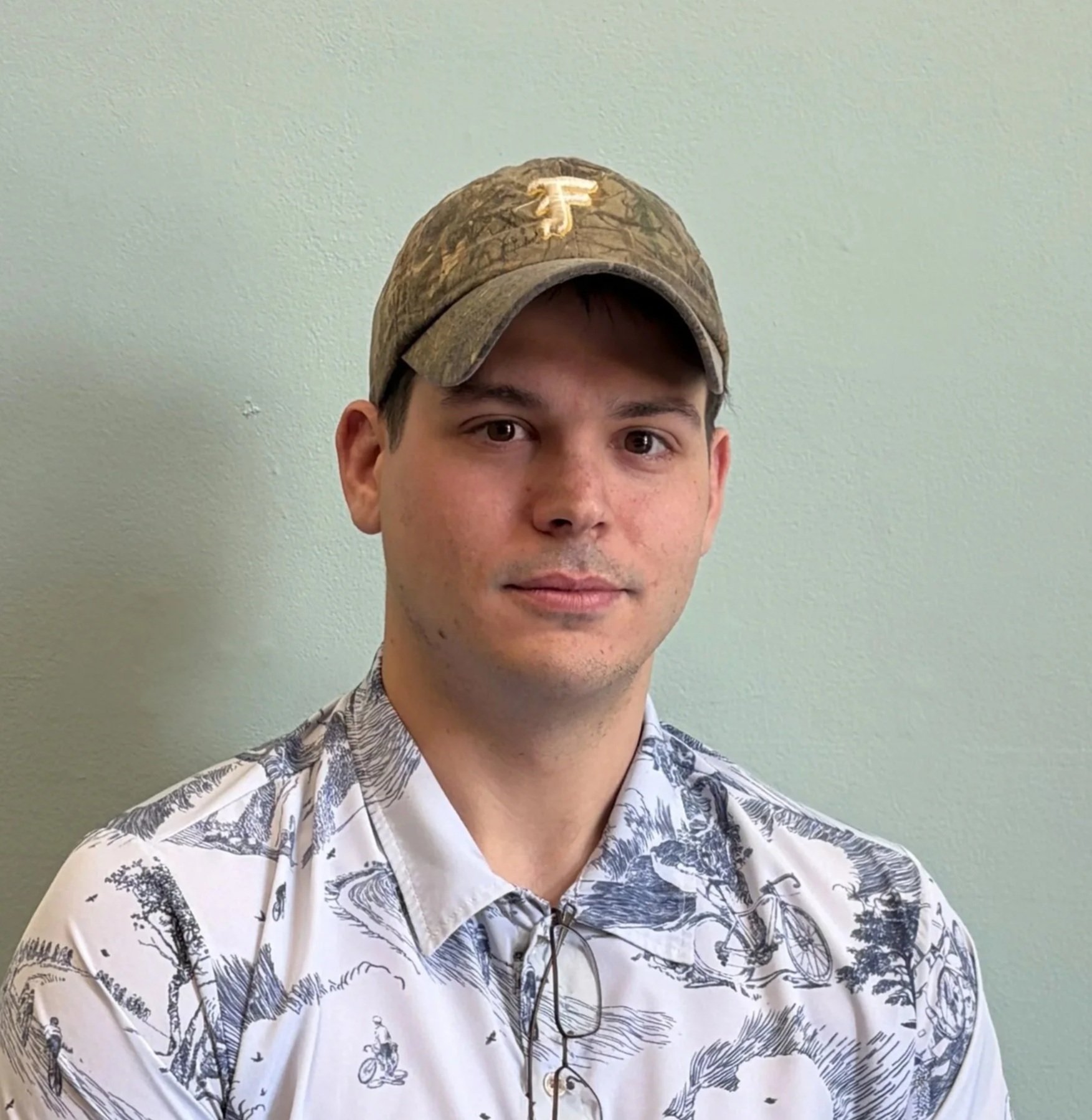 A young man with short dark hair and light skin wearing a camo baseball cap with a gold logo and a white tropical print short-sleeve button-up shirt, standing against a plain light green wall.