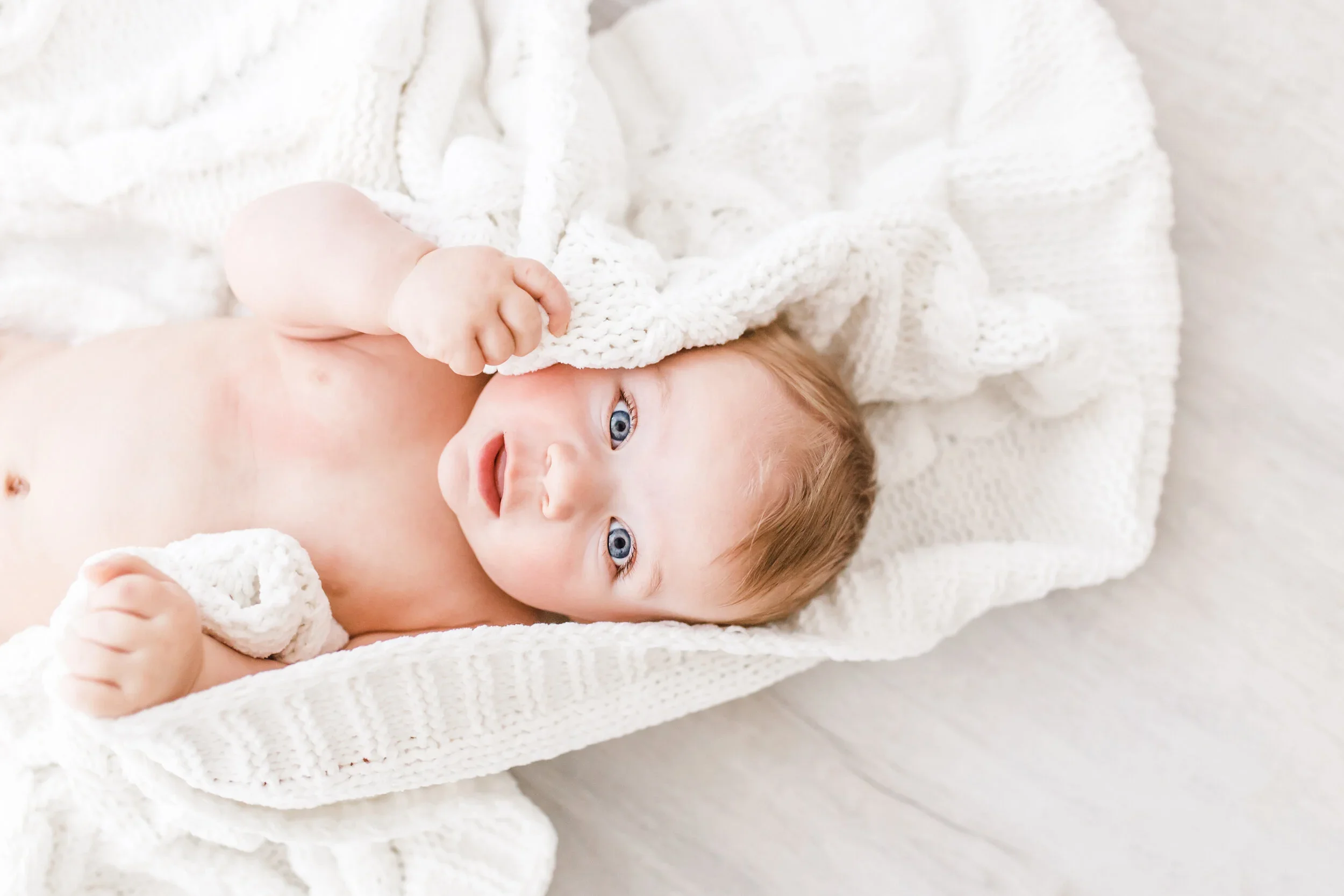 Close-up of a baby with blue eyes lying on a soft white blanket, holding a knitted toy, wearing a cream-colored sweater.