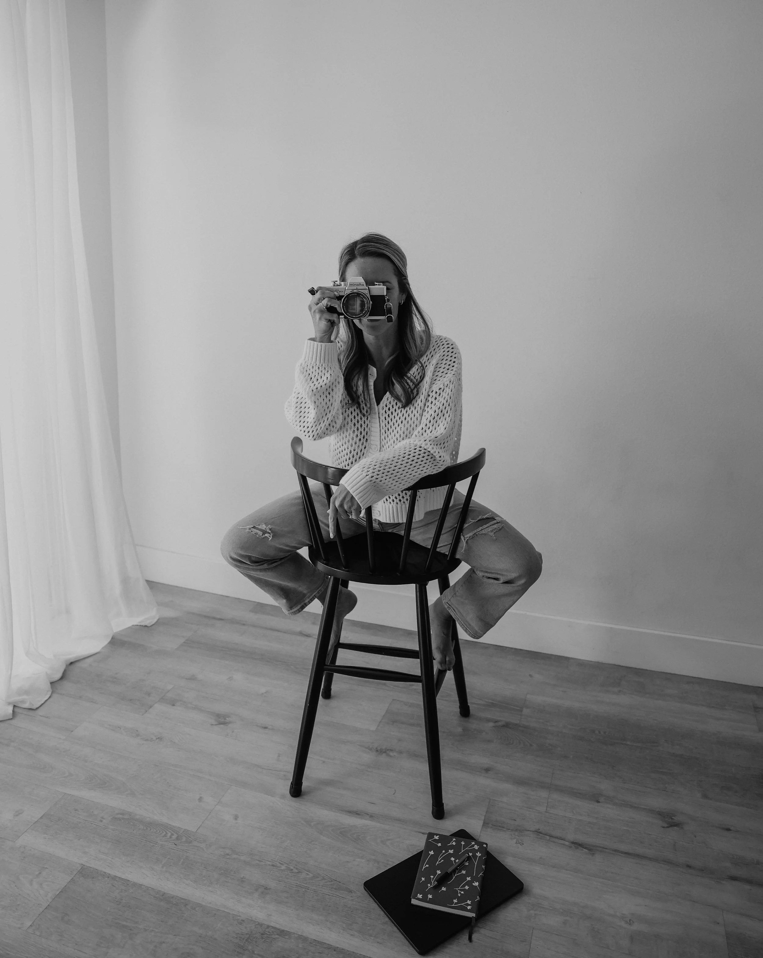 A woman sitting on a chair, taking a photo with a camera, with books and notebooks on the floor beside her, in a room with light-colored walls and wooden flooring.