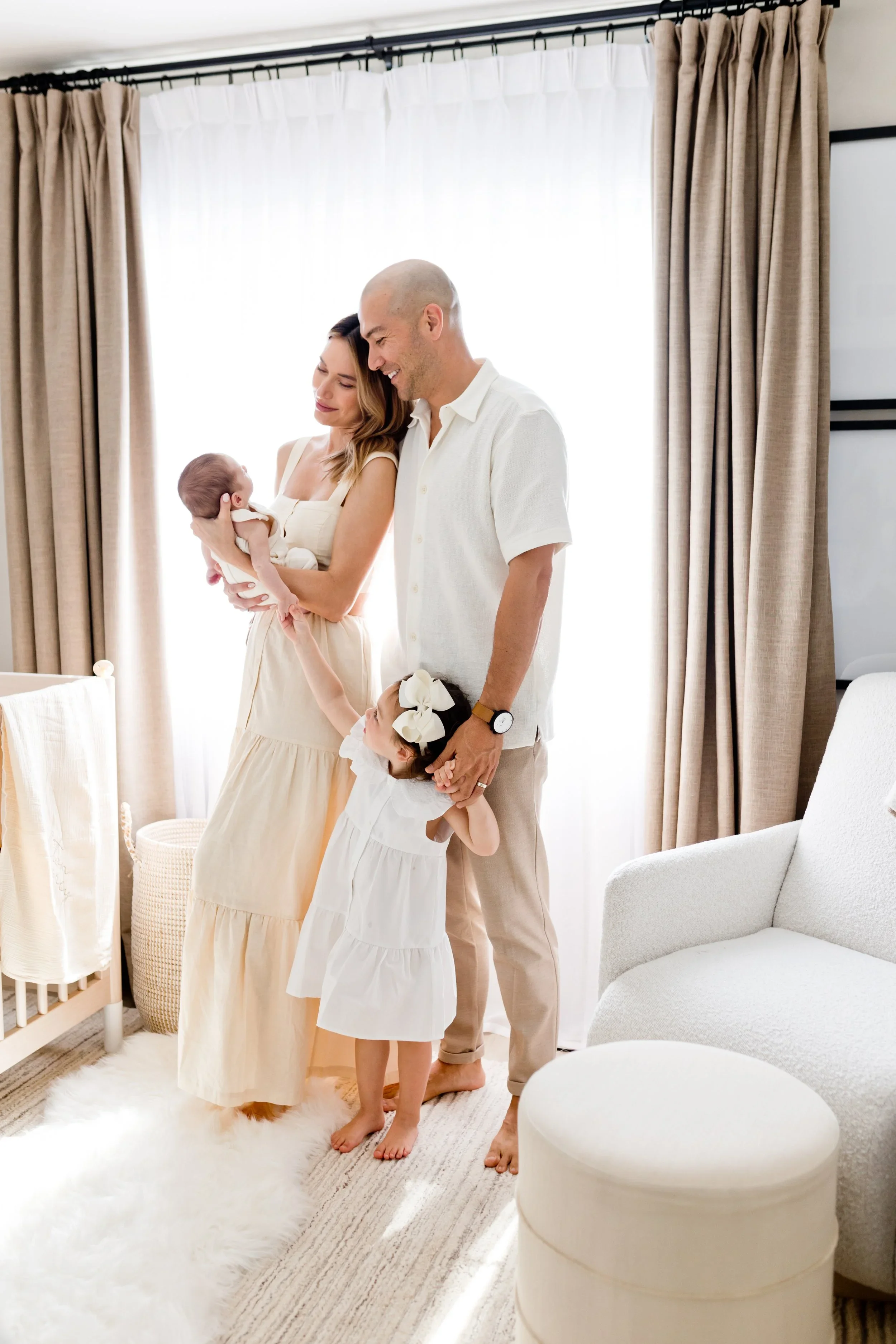 A family in a cozy, well-lit room with beige and white decor. The woman holds a newborn baby, and a young girl with a large white bow in her hair stands nearby, holding her father's hand. The father smiles, and a woman with long hair stands beside him. The room has curtains, a plush white rug, and modern furniture.
