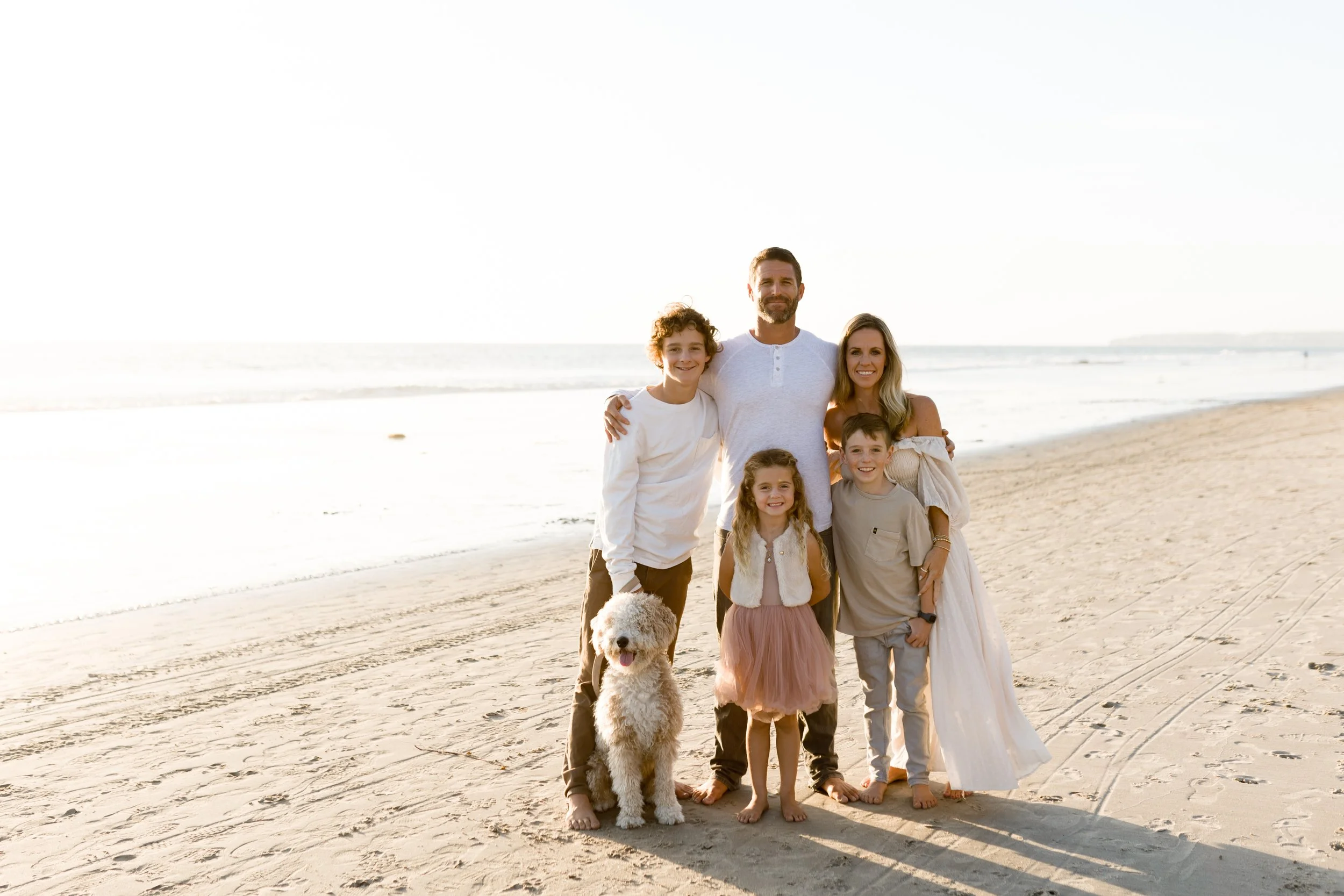 A family of six, including a dog, on a beach, smiling and standing close together with arm around each other.