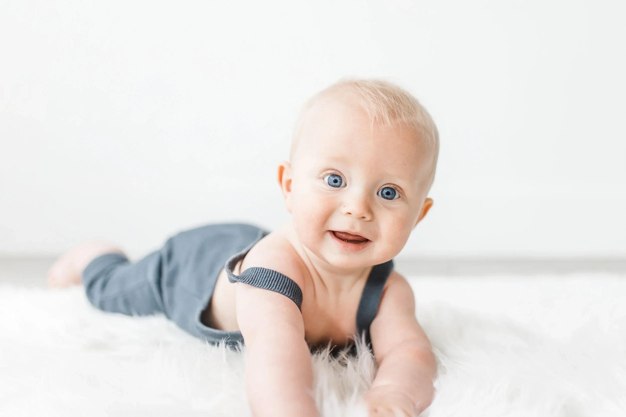 A baby with blue eyes and light blonde hair crawling on a white furry rug, smiling and looking at the camera.