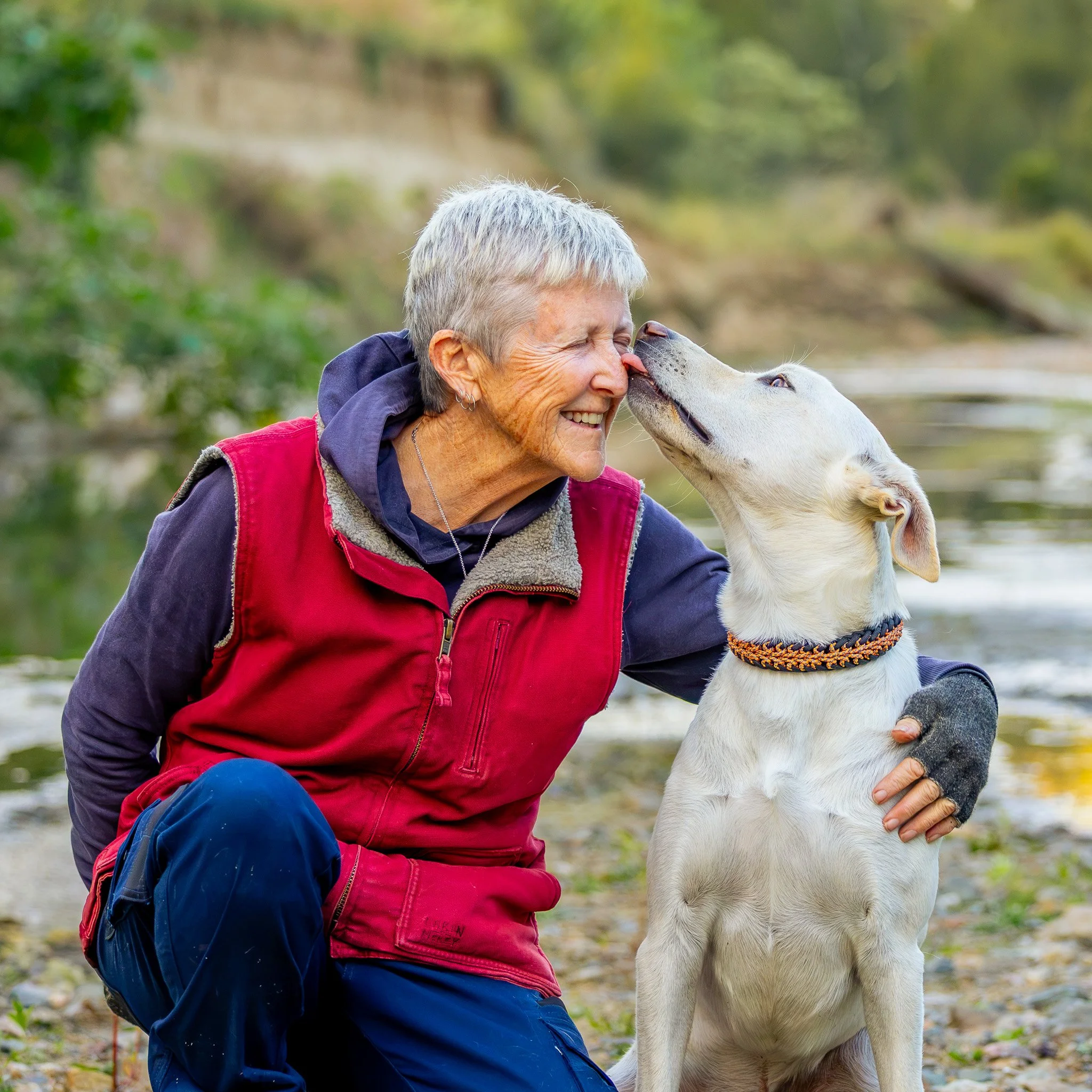 An elderly woman with short gray hair smiling and leaning close to a white dog with light markings, who is touching noses, outdoors with blurred green foliage in the background.  Photographed by Adam Wyatt Photography.