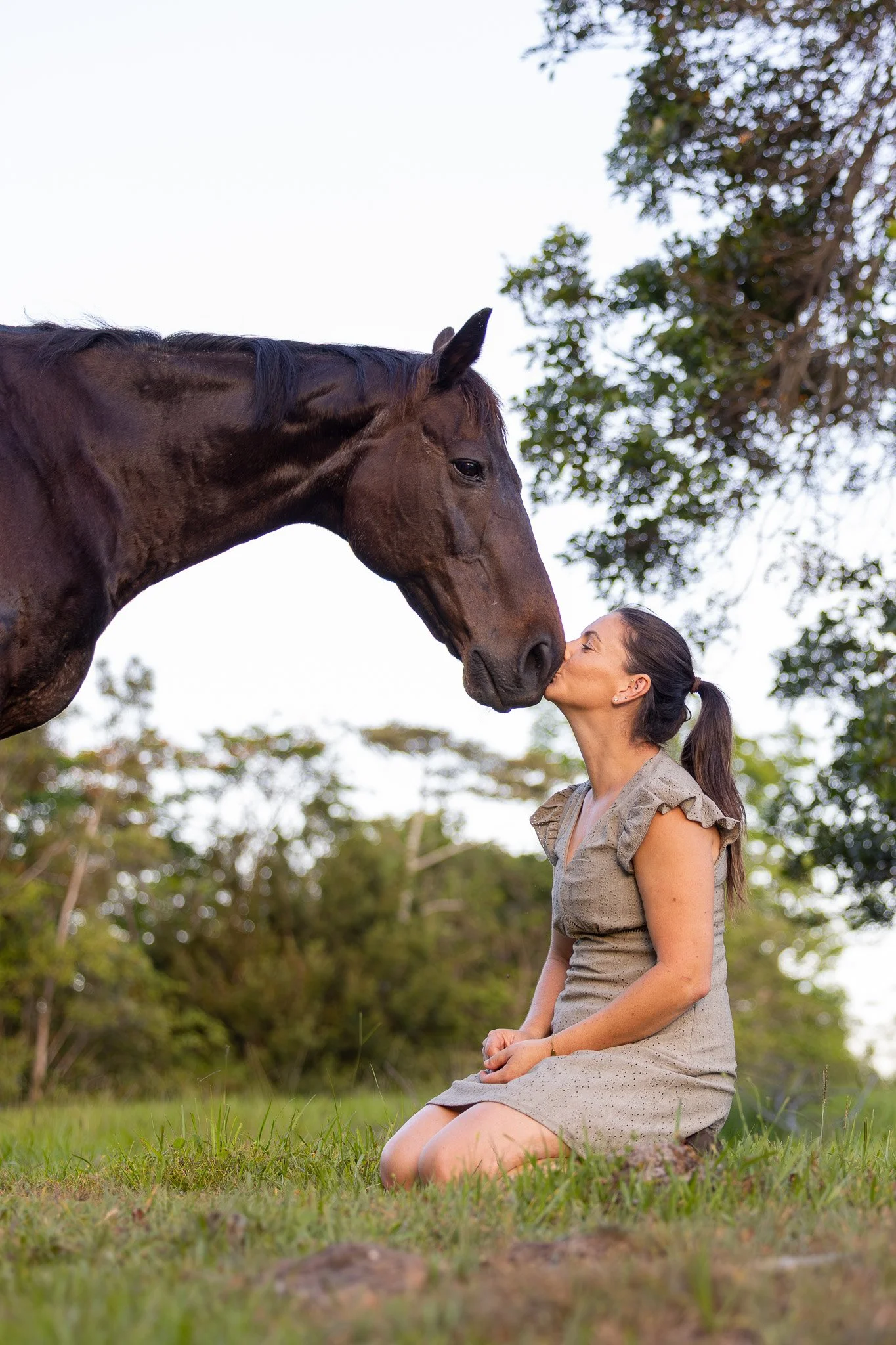 A woman kneeling on grass, kissing a large brown horse on the nose outside during daytime with trees in the background.