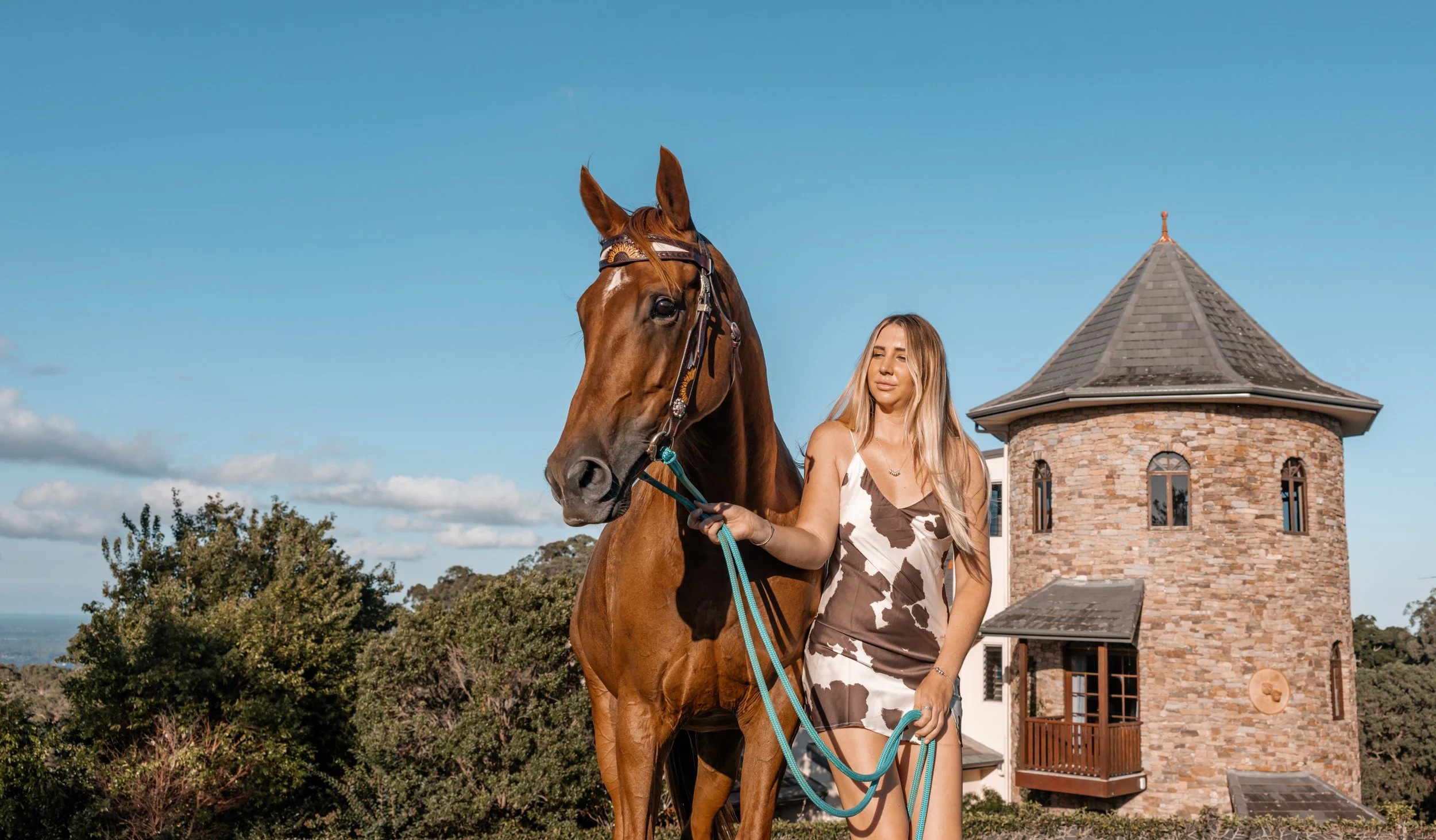A woman with long blonde hair holding a brown horse with a bridle and leash in front of a stone house with a tower. Trees and a blue sky with clouds are in the background.