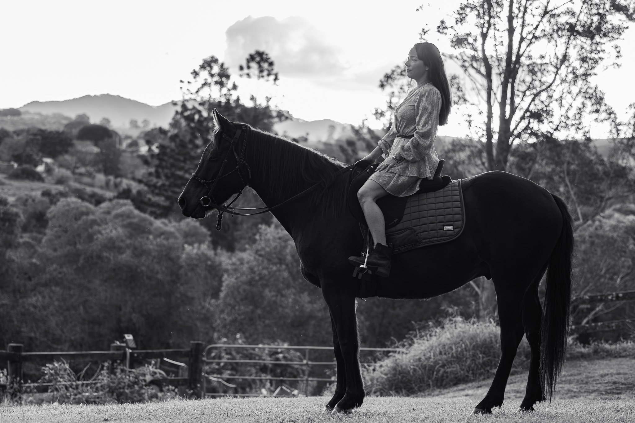 A woman in a dress riding a horse outdoors in a natural setting, with trees and hills in the background. Photographed by Adam Wyatt.