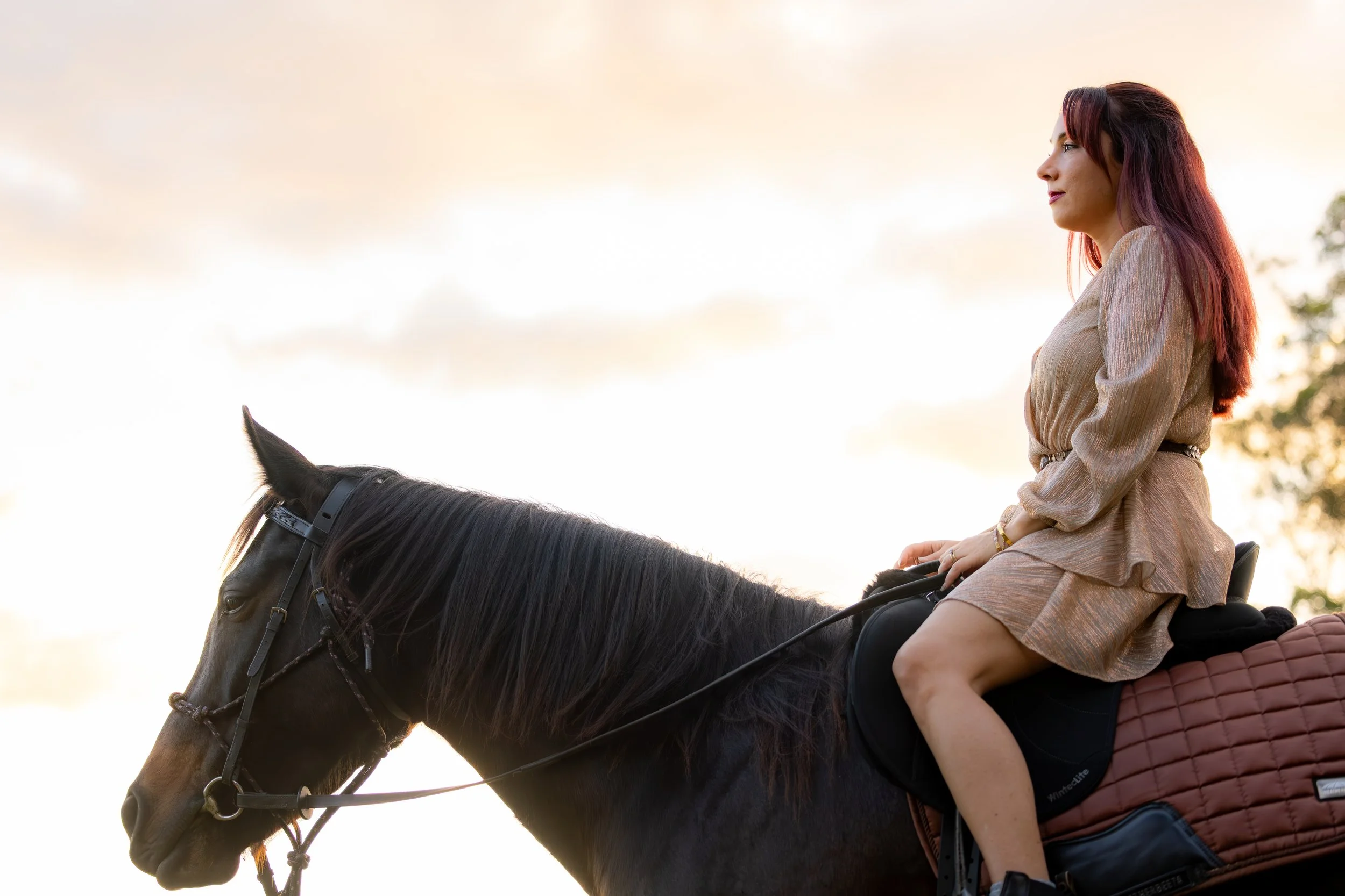 A woman with red and purple hair riding a black horse during sunset. She is wearing a beige dress and sitting upright with her hands on the reins. The sky is partly cloudy with a soft glow from the setting sun.