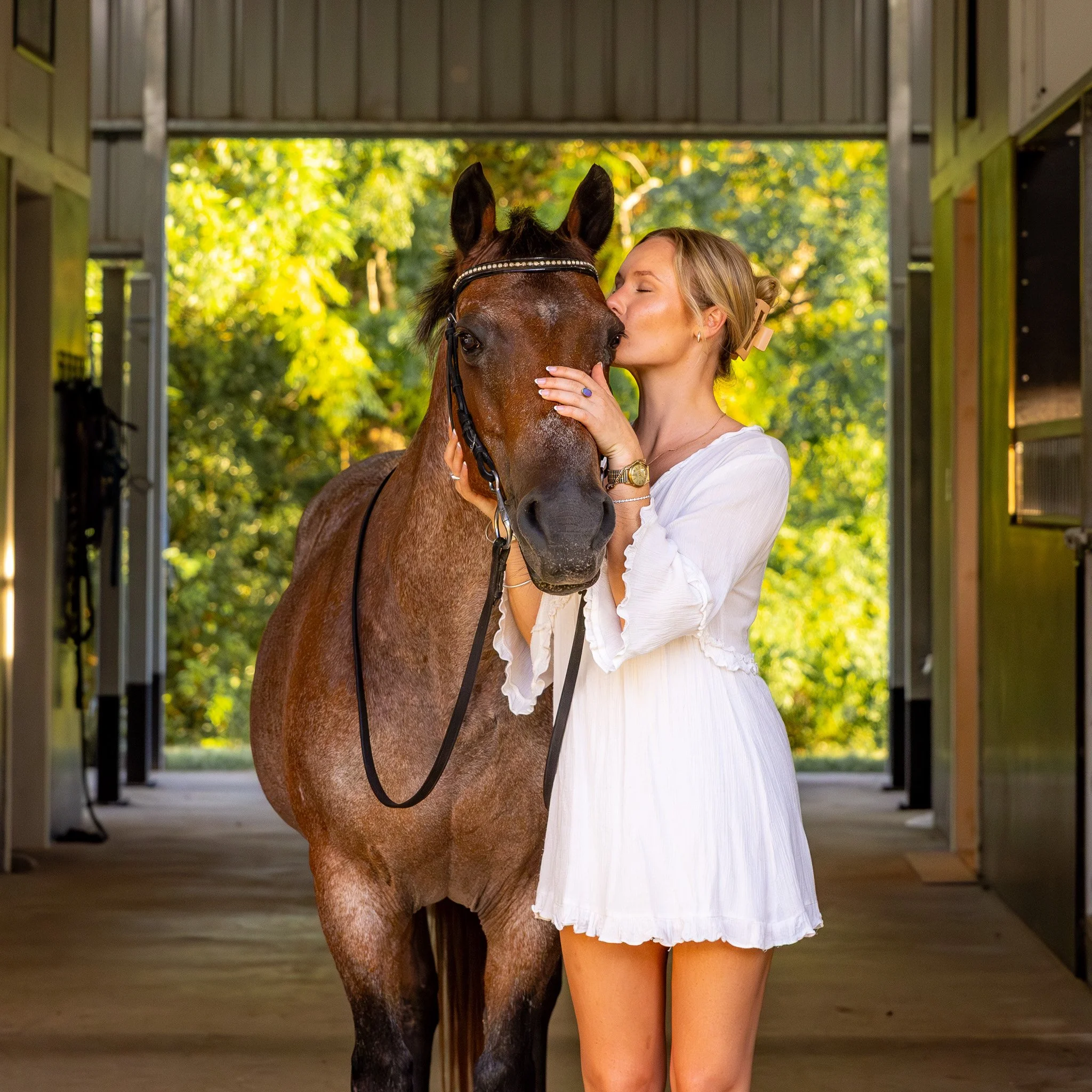 A woman hugging a brown horse under a barn with a background of greenery and trees.