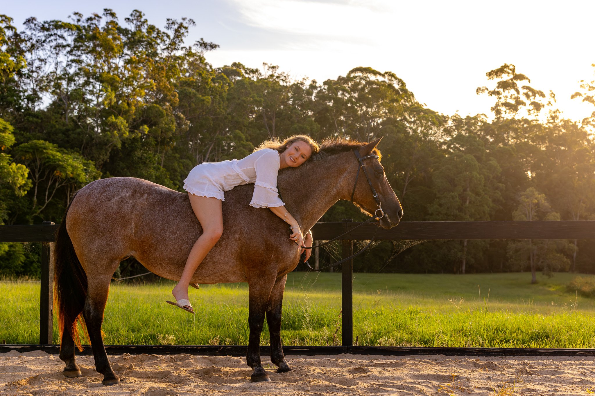 Woman with long hair lying on the back of a brown horse with a black mane, set in a lush green outdoor area with trees and a fence, during sunset.
