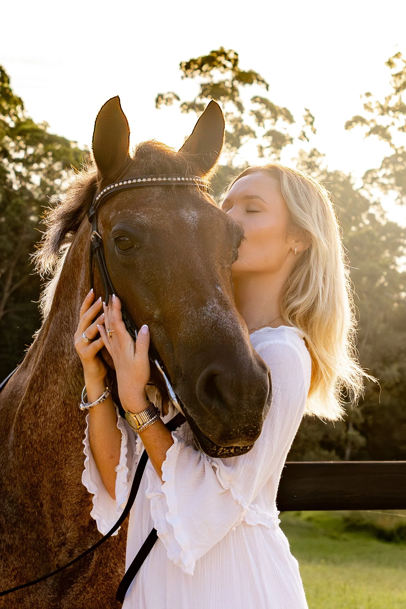 A woman with blonde hair and a white dress kissing a brown horse on the nose outdoors during sunset. Photographed by Adam Wyatt.
