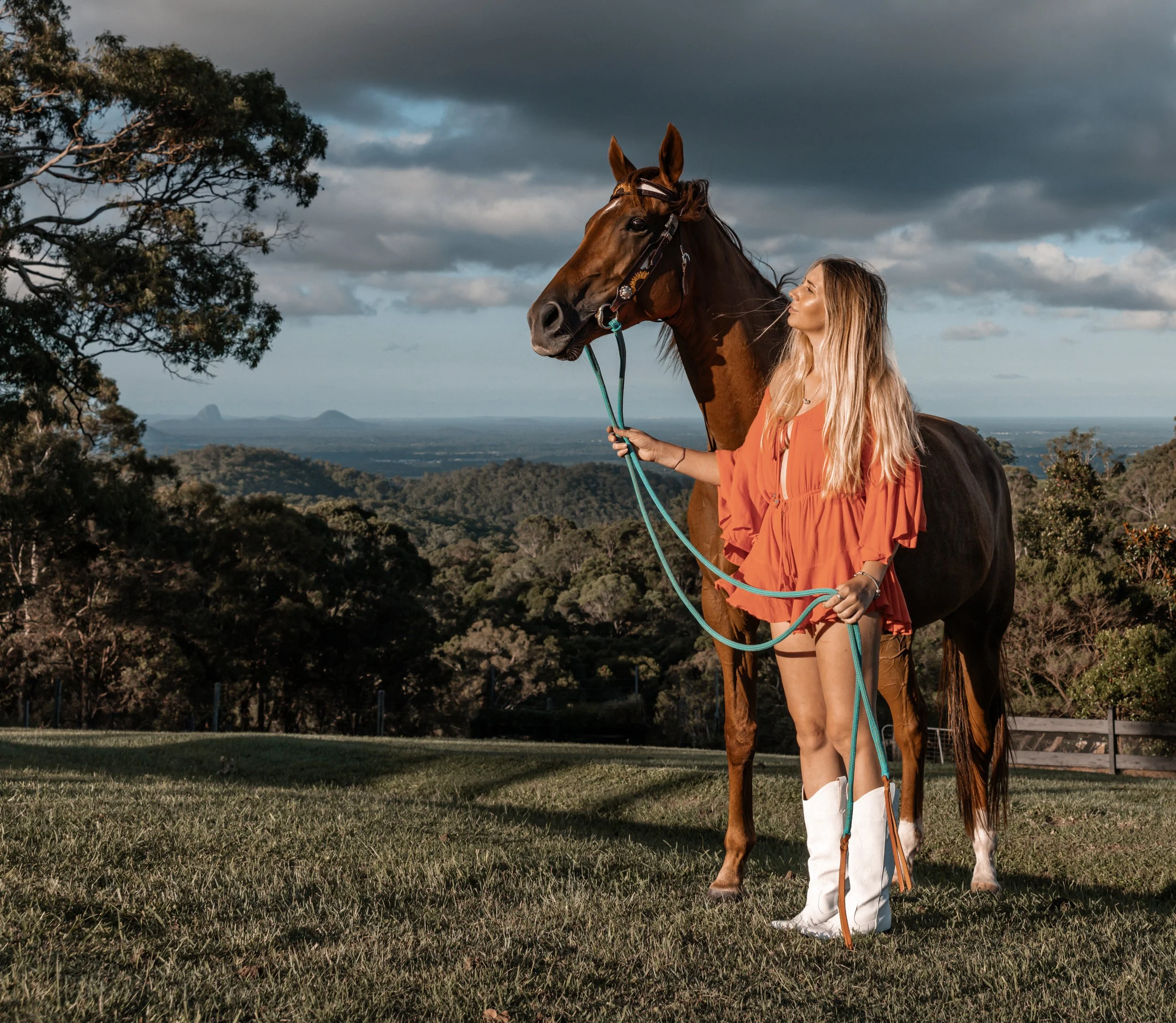 A woman with long blond hair holding the reins of a brown horse outdoors on a grassy field, with trees and a cloudy sky in the background.